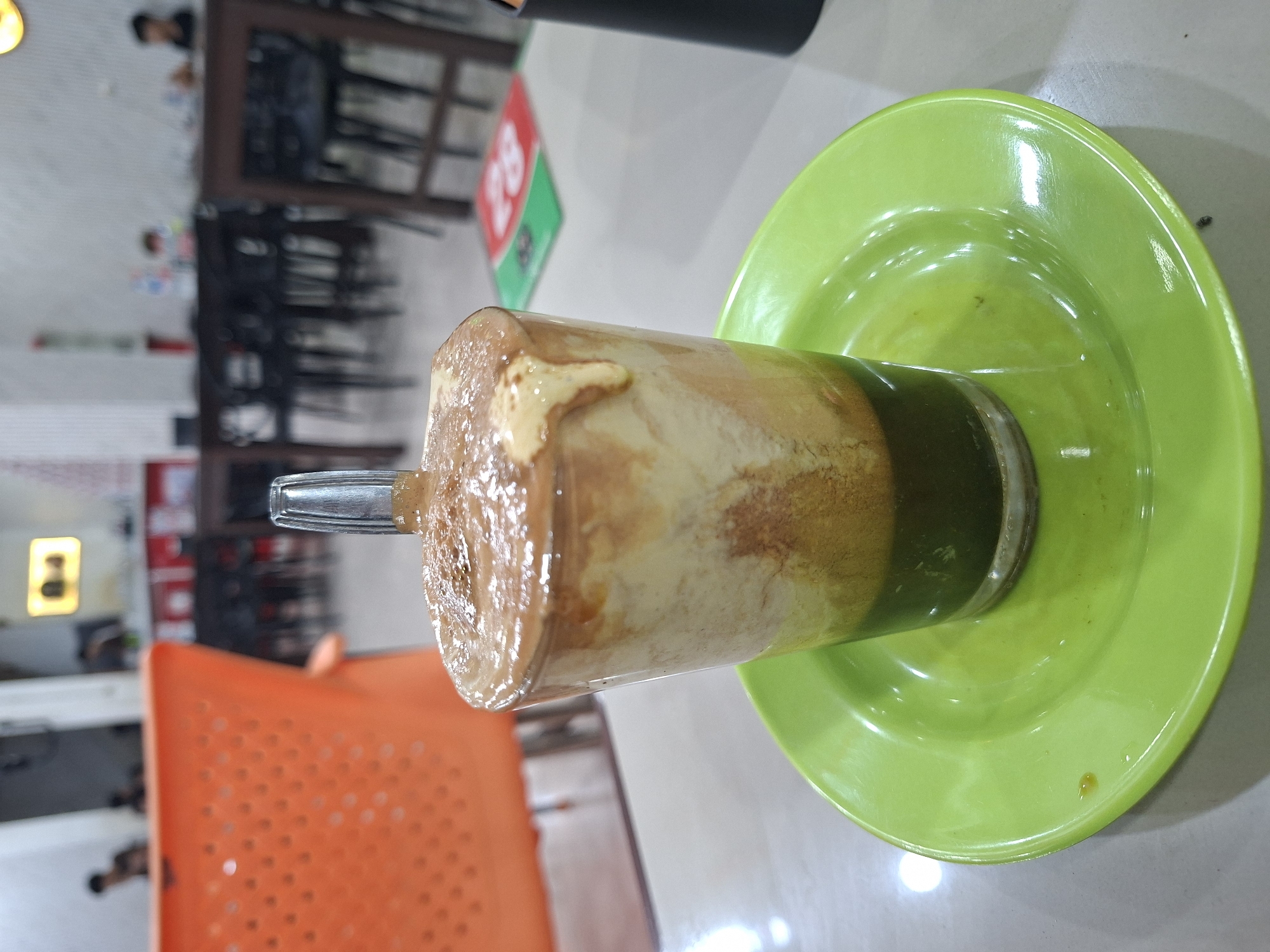 A glass of iced coffee with milk and a green base is placed on a bright green saucer on a white table. The drink has a foamy, light brown top layer, with streaks of white and brown within. The green base is visible at the bottom of the glass. In the background, the blurry interior of what appears to be a cafe or restaurant is visible. Orange chairs and dark wooden tables with chairs are present. Faint figures of people can be seen in the background. A red and green floor mat with the number "28" is visible in the middle ground. The overall lighting suggests an indoor environment, possibly during the daytime. This scene is likely located in the city of Sigli, Indonesia, given the context provided.
