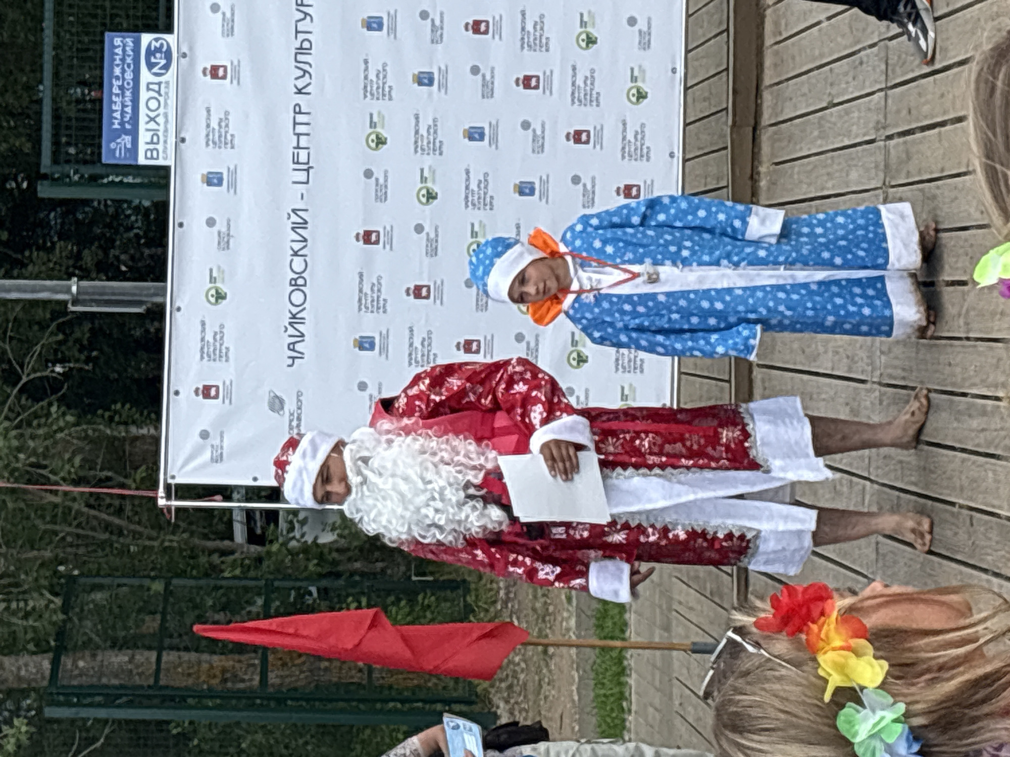 In Chaikovsky, Russia, two individuals dressed in festive attire stand on a wooden platform. The person on the left is dressed as Ded Moroz, a Russian Santa Claus figure, wearing a red velvet coat with intricate white patterns, a white beard, and a red hat. They are holding a piece of white paper. To their right stands a child in a blue coat with white polka dots, a matching blue hat with a white pom-pom, and an orange-lined hood. The child also wears a medal around their neck. Behind them is a large banner with text in Russian, including "ЧАЙКОВСКИЙ - ЦЕНТР КУЛЬТУРЫ" (Chaikovsky - Center of Culture) and various smaller logos. A red flag on a pole is visible to the left of Ded Moroz. In the foreground, the back of a person's head with a colorful flower crown is visible. The environment appears to be outdoors, with trees and a green fence in the background.
