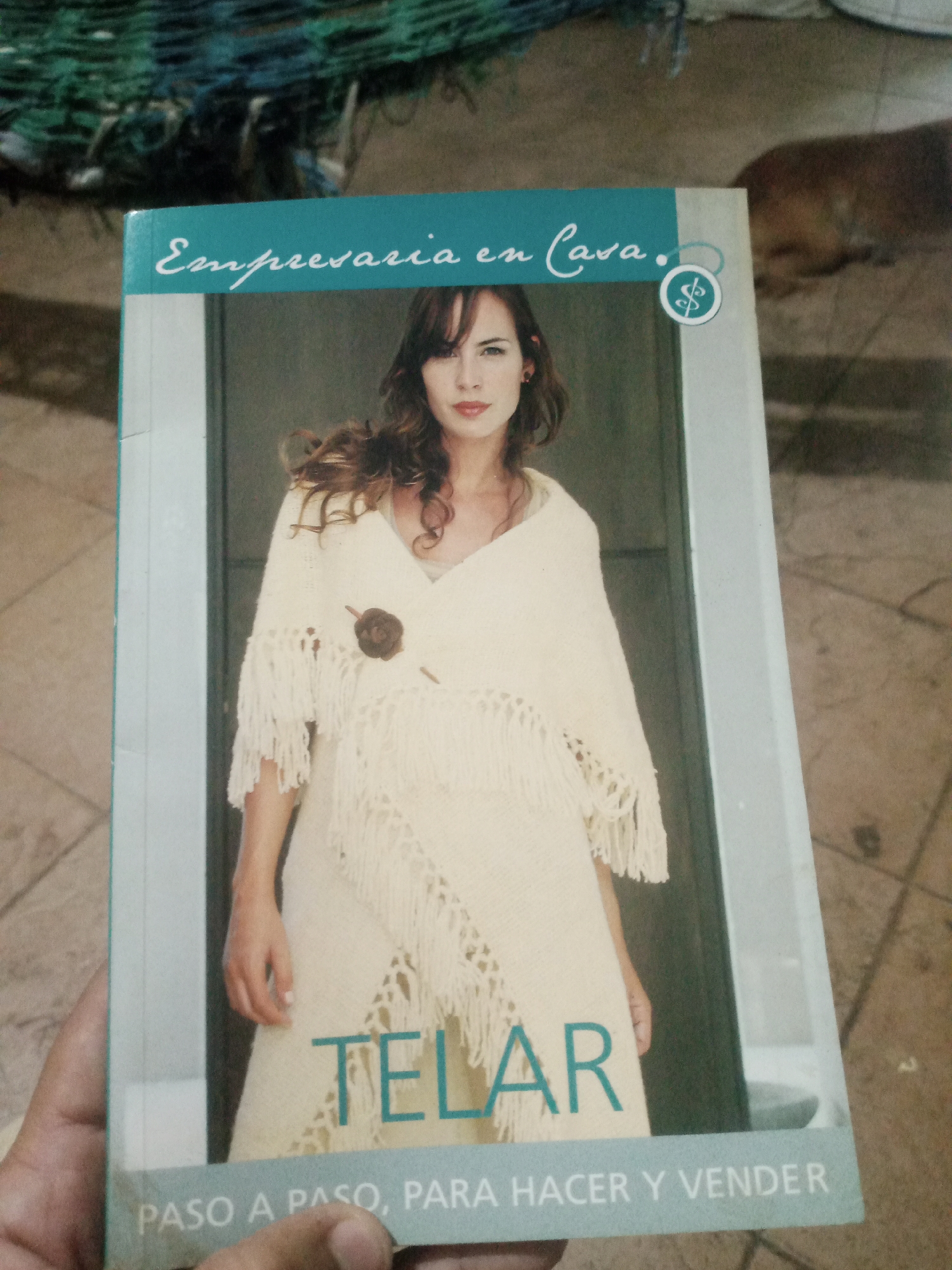 The image shows a close-up of a book cover titled "Empresaria en Casa. Telar." The cover features a woman wearing a cream-colored knitted shawl with fringe. The title is in Spanish, with "Telar" being a prominent word in large, teal-colored letters. Below this, the phrase "PASO A PASO, PARA HACER Y VENDER" is visible. A hand is holding the bottom of the book. The background is out of focus, but appears to be an outdoor setting with textured ground and some green netting at the top. The book cover itself has a light blue border. There is a small dollar sign symbol enclosed in a circle on the top right of the cover.