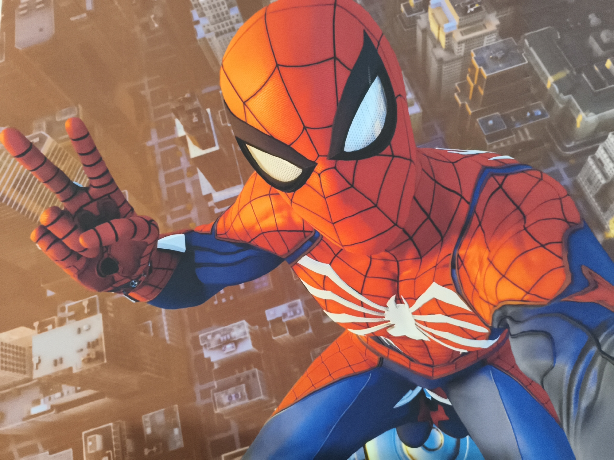 A full shot from a slightly low angle captures a costumed character resembling Spider-Man, posed with their left hand held up in a peace sign. The character is predominantly red with blue accents and a white spider emblem on the chest. The background is a blurred cityscape, suggesting an urban environment. The lighting is warm and appears to be from an overhead source, casting subtle shadows and highlighting the texture of the costume. The character's eyes, visible through the mask, are white and appear to be lenses.
