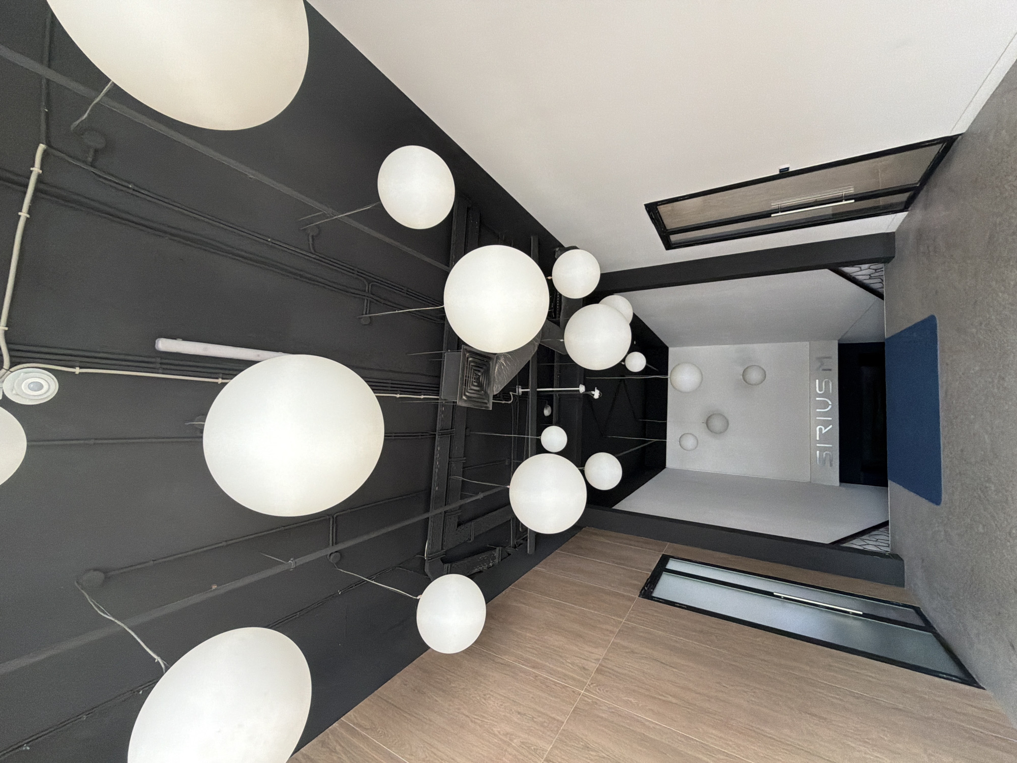 This media file is an upward-looking photograph of a modern interior space, likely a public building or office. The scene is dominated by a dark, industrial-style ceiling with visible black conduits and pipes. Suspended from the ceiling are numerous large, white, spherical pendant lights of varying sizes, creating a striking visual element. The lights are distributed unevenly throughout the upper portion of the frame.
Below the dark ceiling, the environment transitions to a brightly lit, minimalist interior with white walls and a light-colored floor that appears to be tiled with a wood-grain pattern. There are dark structural elements and some glass-fronted openings visible, suggesting a multi-level or enclosed space. A blue mat is visible on the floor in the far background.
On one of the white walls, the word "SIRIUS" is displayed vertically in illuminated, blocky, silver lettering. There are also several smaller, circular light fixtures or decorative elements on the same wall. The overall impression is one of contemporary design, with a contrast between the raw, dark ceiling and the clean, bright interior spaces. There are no people visible in the image. The lighting suggests it is daytime, likely indoors, and the weather is not discernible. The location cues are limited to the visible interior architecture and the text, with the provided context indicating Minsk, Belarus.