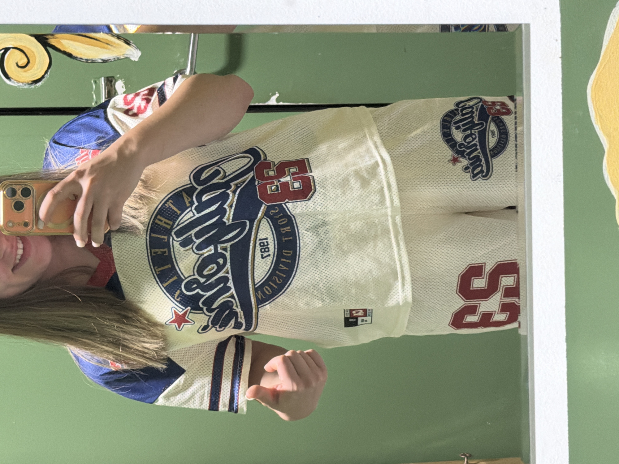 A person is reflected in a mirror, holding a mobile phone and taking a selfie. They are wearing a baseball jersey with "California Angels 1983 Division" and the number "53" prominently displayed. The mirror is framed and mounted against a pale green wall with additional artistic elements. The setting appears to be indoors. There is no information in the image to place the location within Arroyo de la Miel-Benalmádena Costa, Spain.