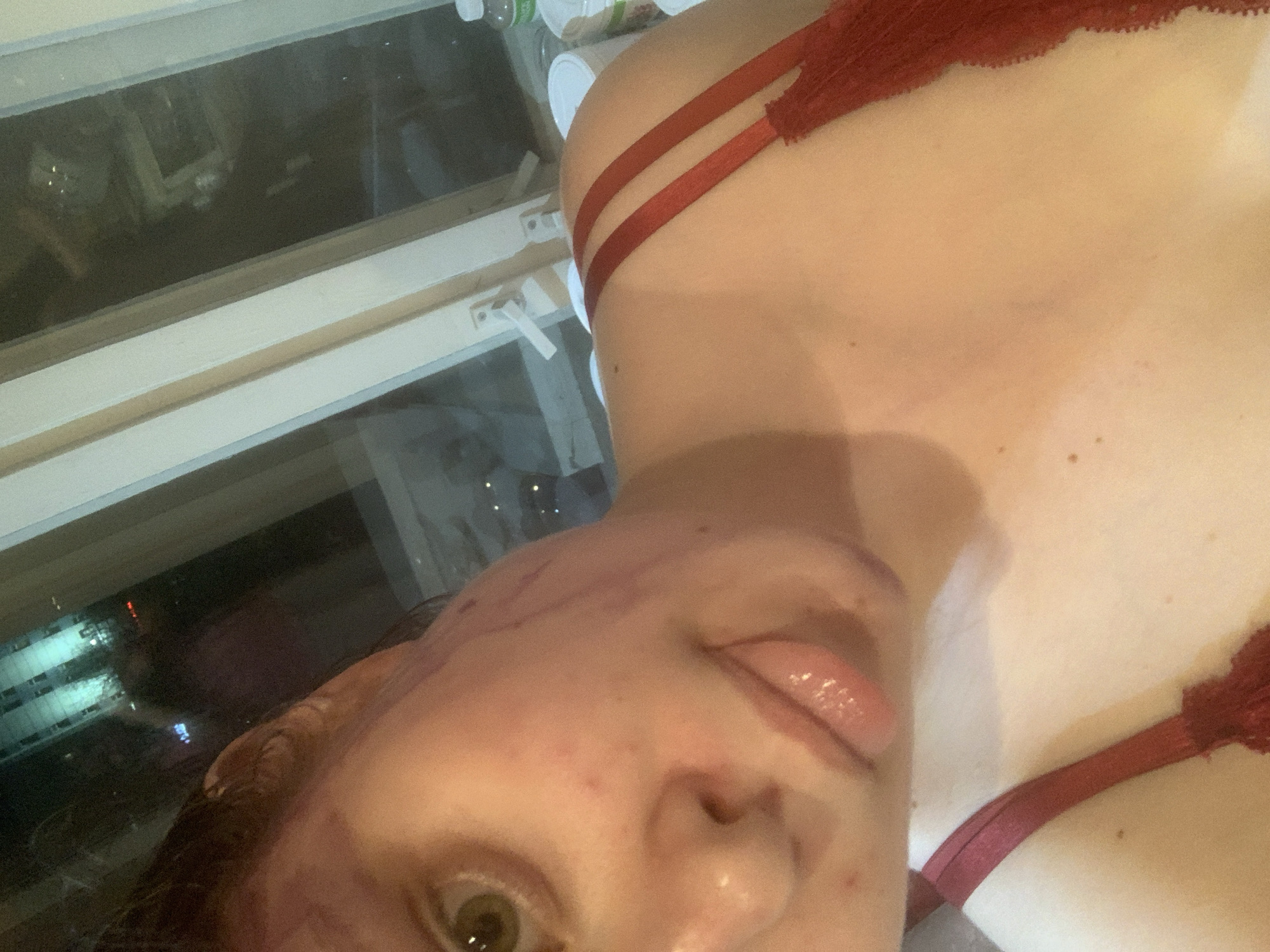A close-up, angled selfie captures a person indoors at night. The subject, likely female, is partially visible, featuring one eye (greenish-brown), nose, and mouth with the tongue slightly extended. There are prominent reddish-purple marks or smudges on the person's cheek and forehead. They are wearing a red bra or lingerie with two visible straps over the shoulder.
In the background, visible through or reflected in a window, are several plastic bottles and white containers, possibly food-related. Beyond the window, a dark nighttime cityscape or residential area is illuminated by artificial lights from buildings. No specific activity beyond posing for the selfie is evident. There is no visible text in the image.