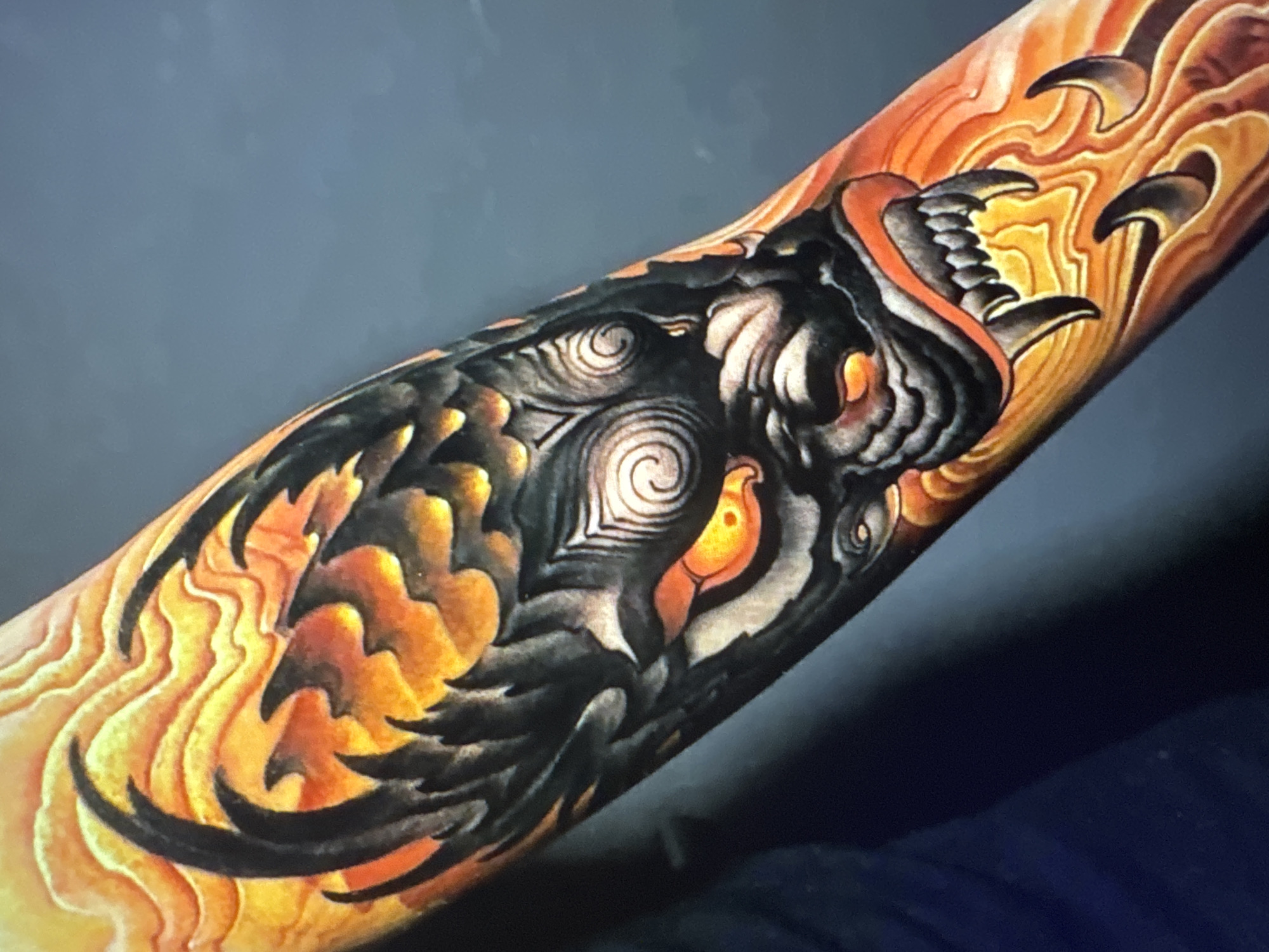 A close-up, eye-level, medium shot captures a detailed tattoo of a fierce dragon or demon face on a person's forearm. The tattoo is rendered in vibrant oranges, yellows, and black, with intricate shading and sharp lines. The creature's eyes glow with an intense orange light, and its mouth is open, revealing sharp fangs. Flames, depicted as swirling patterns of orange and yellow, surround the face. The background is a solid, dark gray, suggesting an indoor setting or a dimly lit area. The skin texture and the tattoo's details are sharply in focus.