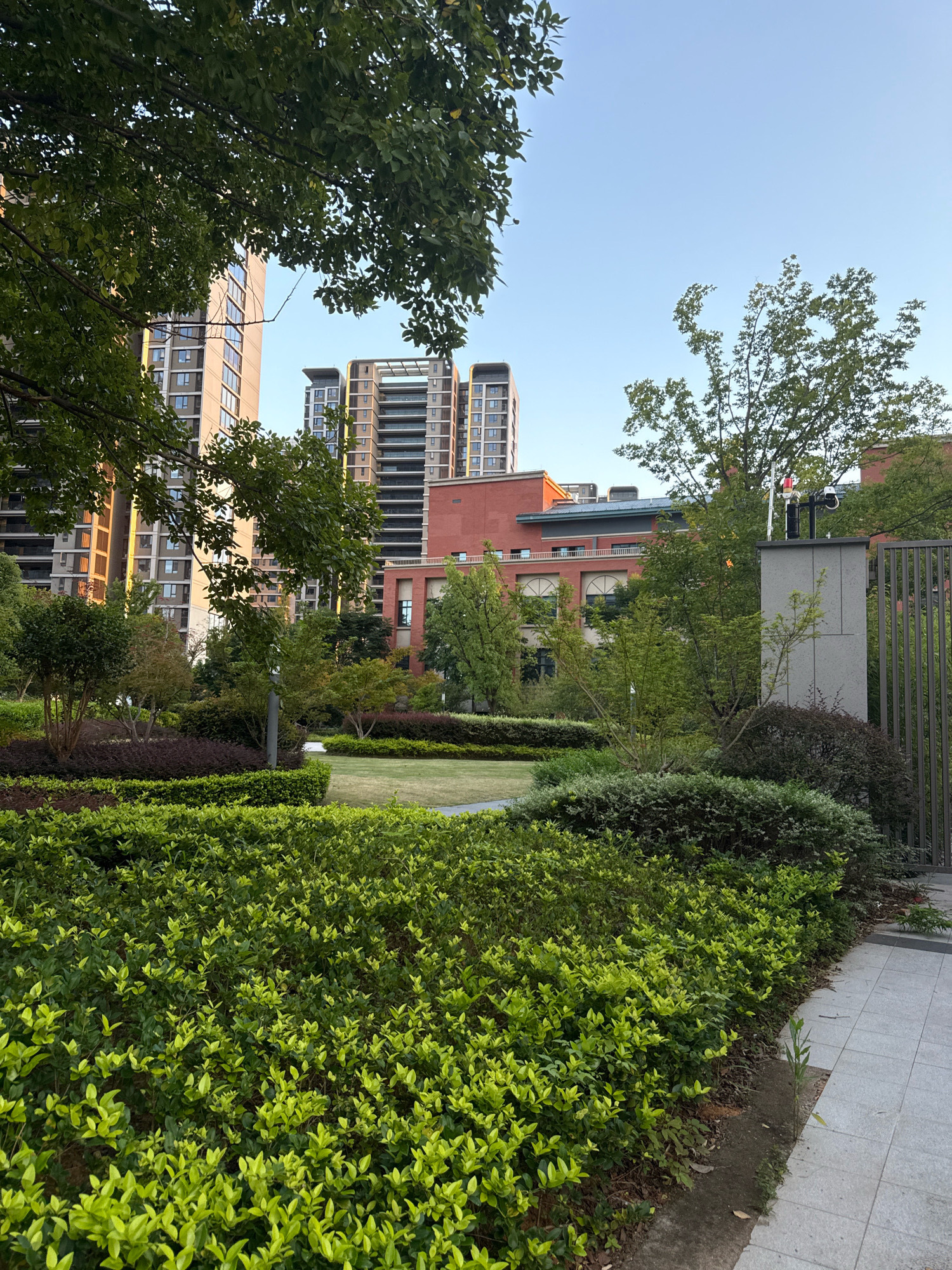 The image depicts a modern urban landscape with lush greenery. In the foreground, a meticulously manicured hedge of bright green shrubs lines a paved walkway. Behind this, several layers of well-maintained bushes and small trees form a verdant barrier, with a neatly trimmed lawn visible in the mid-ground. A grey concrete pillar topped with a security camera and red and white warning lights stands to the right, adjacent to a vertical metal fence.
In the background, a complex of contemporary buildings rises. The structures feature a mix of beige, brown, and red brick facades, with numerous windows. The architectural style suggests residential or commercial high-rises. Trees with green foliage are interspersed throughout the scene, softening the urban architecture. The sky above is a clear, pale blue, indicating fair weather. The overall impression is one of a well-designed, contemporary residential or business complex in a Chinese city. The specific location of Yiwu, China, can be inferred from the context of the provided image.