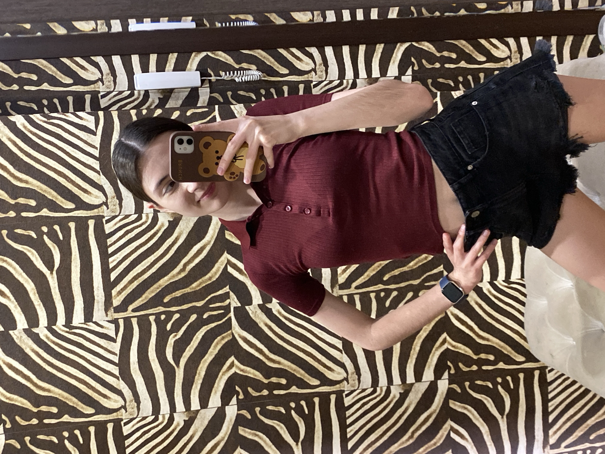 The image shows a young woman taking a selfie in front of a wall with a zebra print pattern. She is wearing a maroon polo shirt and black denim shorts with a frayed hem. She is holding a phone with a brown bear-themed case in her left hand and has a smartwatch on her left wrist. The location appears to be indoors. The context of being in Votkinsk, Russia is not verifiable from the image.
