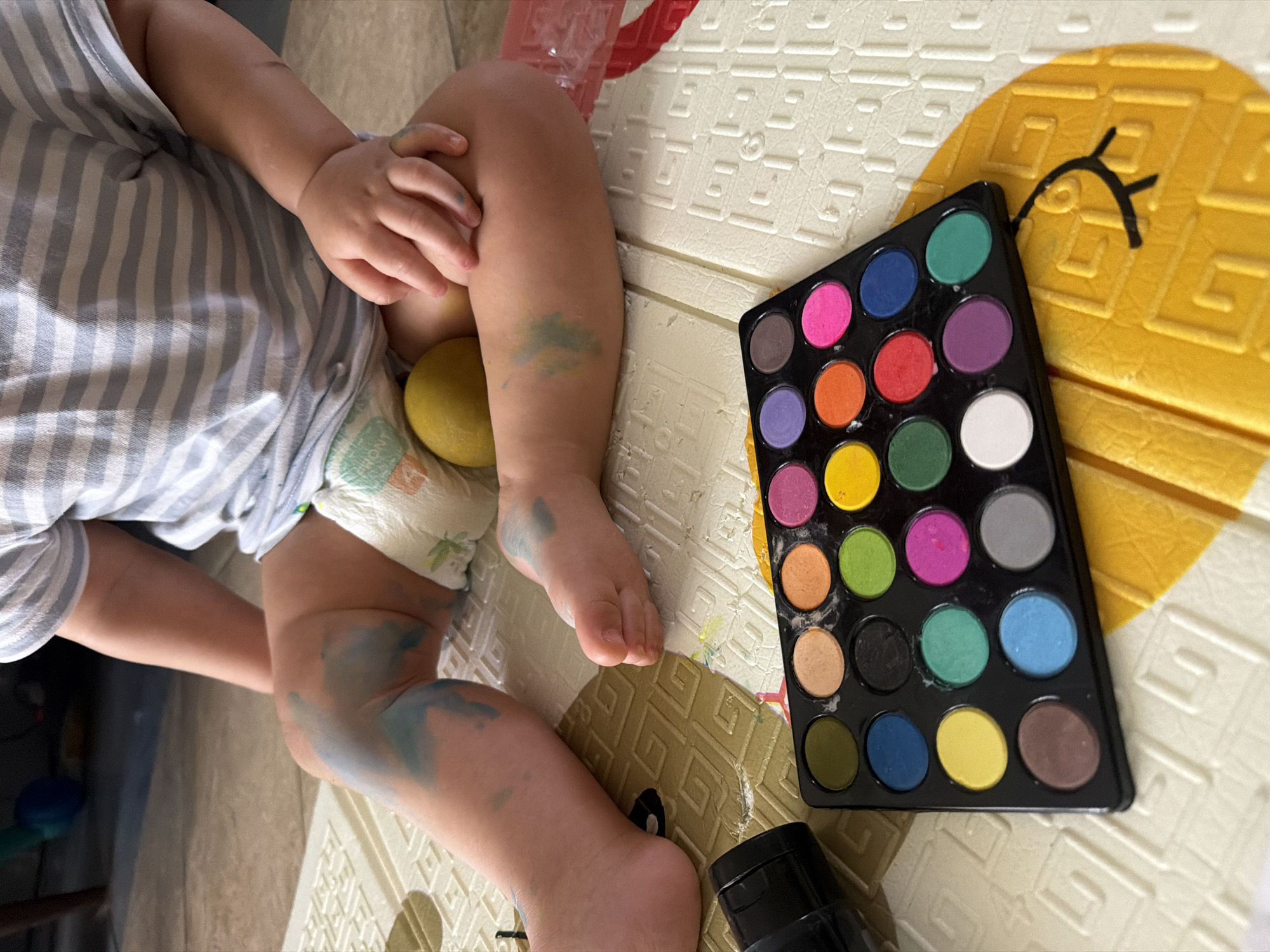 A toddler, wearing a striped shirt and a diaper, is sitting on a yellow and white playmat with blue paint smudges on their legs. A round, yellow object, possibly a lemon, is nestled between their legs. In front of the toddler is a black palette with 24 circular pans of watercolors, some of which have been used. A black object, possibly a tube of paint or a brush, is visible to the left of the palette. The playmat has a patterned texture and a cartoon-like drawing of a face on it. The background is blurred, showing a tiled floor and furniture. The image appears to be taken indoors, likely in a residential setting. There is no visual information within the image that indicates its location in Sharm El Sheikh, Egypt.