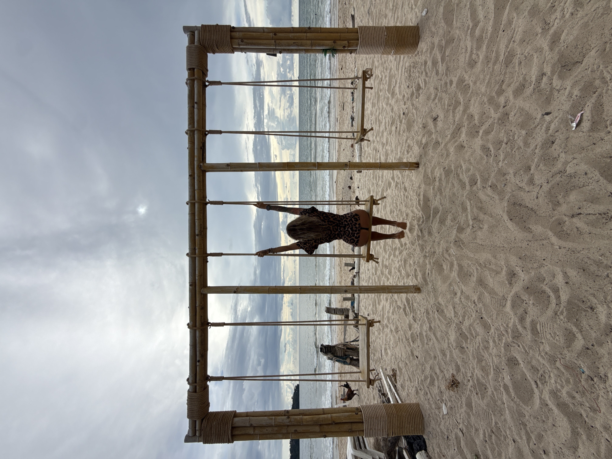 A woman with long blonde hair, wearing a leopard-print dress, sits on a wooden swing at a beach in Choeng Thale, Thailand. She is facing away from the camera, with her arms raised and gripping the ropes of the swing. The swing set, constructed from bamboo and rope, is positioned on a sandy beach facing the ocean. The sea is calm with gentle waves, and the sky is overcast with clouds. In the background, there are some discarded wooden items and a chicken walking on the sand. The overall scene depicts a relaxed, tropical atmosphere.