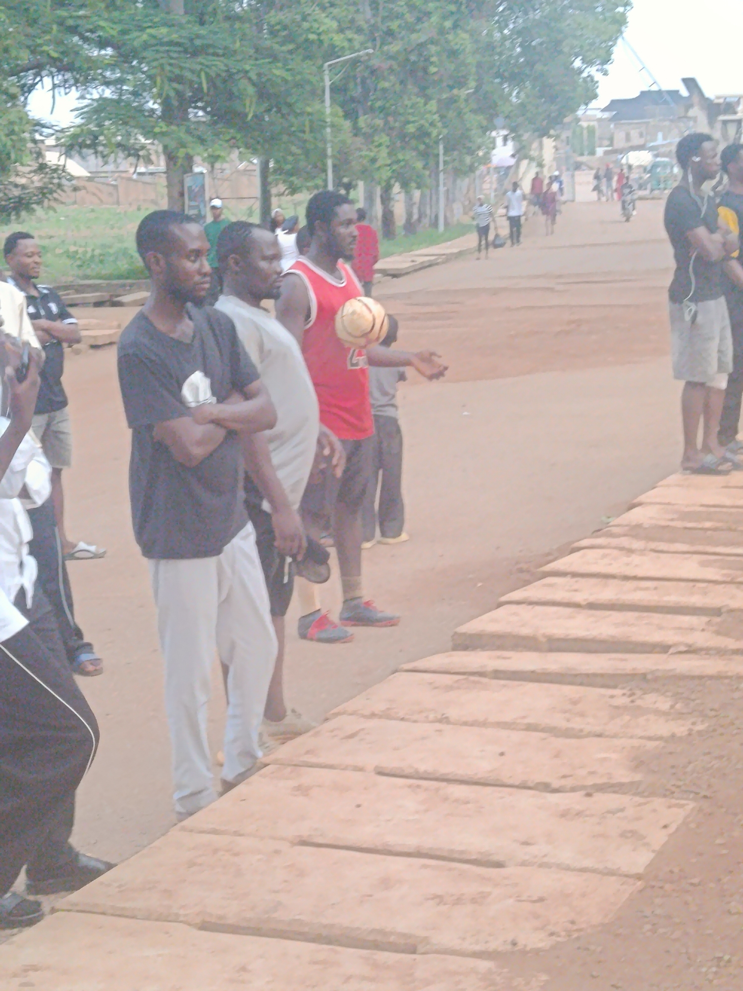 The image depicts a street scene, likely in Rafin Par, Nigeria, with several men standing around. In the foreground, a man wearing a black t-shirt and light grey pants is visible from the chest down, with his arms crossed. To his right, another man in a light-colored t-shirt and grey pants is partially obscured. Further to the right, a man in a red sleeveless shirt is holding a ball. Behind them, a group of people are walking or standing on the dirt road. Trees line the left side of the road, and buildings are visible in the background. The ground is a mixture of dirt and what appear to be raised concrete slabs in the foreground. The overall lighting suggests it is daytime.