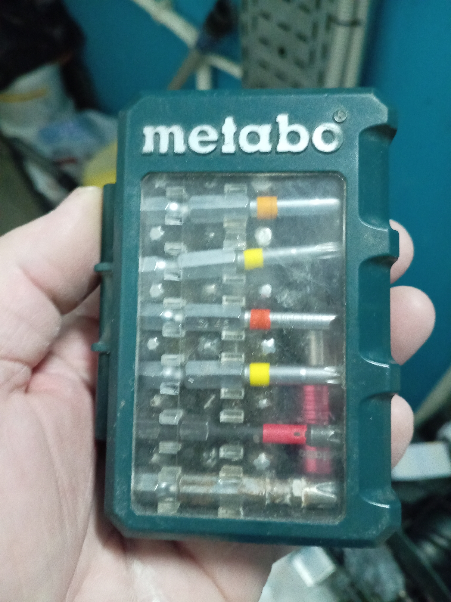 A hand holds a green Metabo case filled with screwdriver bits. The bits are silver with colored bands of orange, yellow, and red. The case is slightly dusty. The location is unknown.