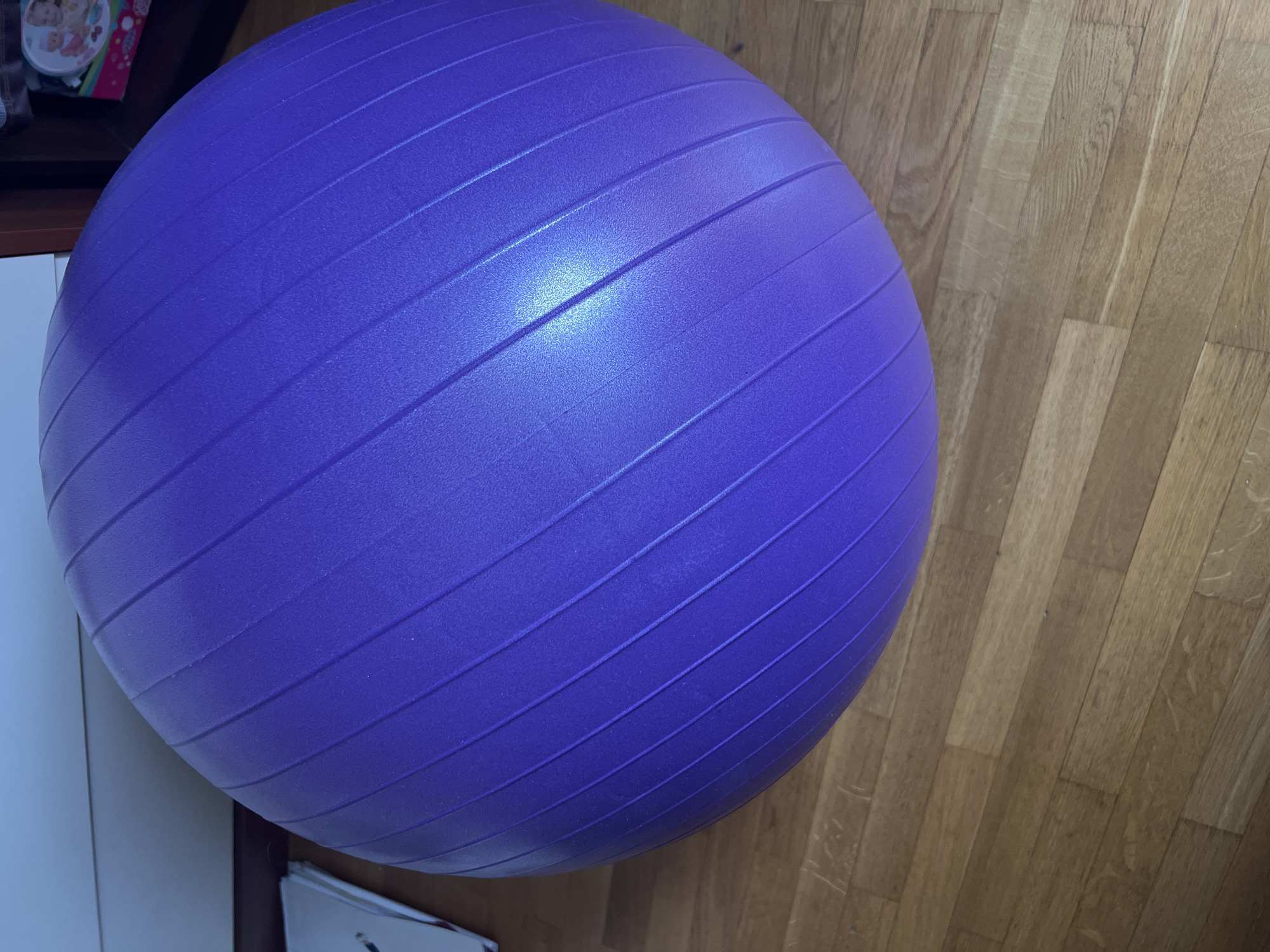 A large, purple exercise ball rests on a polished wooden floor in a room. The ball has a textured surface with raised, parallel lines running diagonally across it. Light reflects off the ball, creating a bright highlight on its upper surface. In the background, the edge of a white surface and a dark wooden cabinet are visible. The scene appears to be indoors, likely in a home or fitness studio, and there is no information to suggest a specific location within Minsk, Belarus. There are no people, actions, or interactions depicted.
