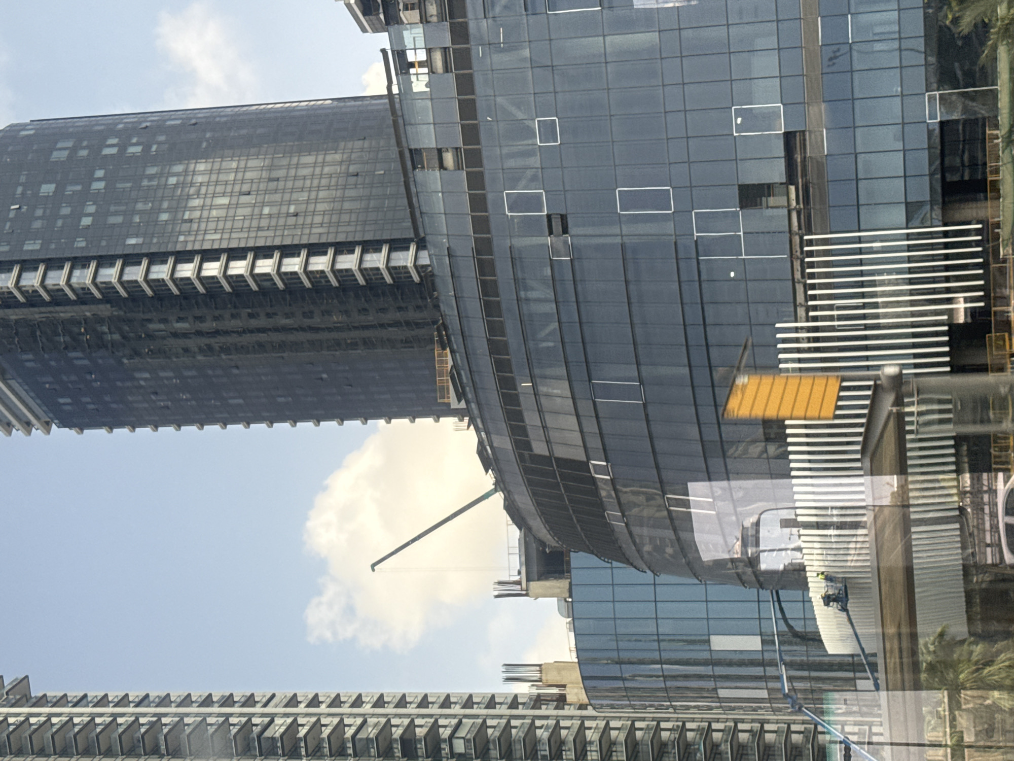 The image shows an upward view of modern buildings under construction in Tel-Aviv, Israel. The structures are primarily made of glass and steel, featuring rectangular patterns and curvilinear designs. Construction cranes are visible against a sky with scattered clouds. Construction equipment and personnel can be seen on scaffolding. Reflections and shadows play across the building surfaces.