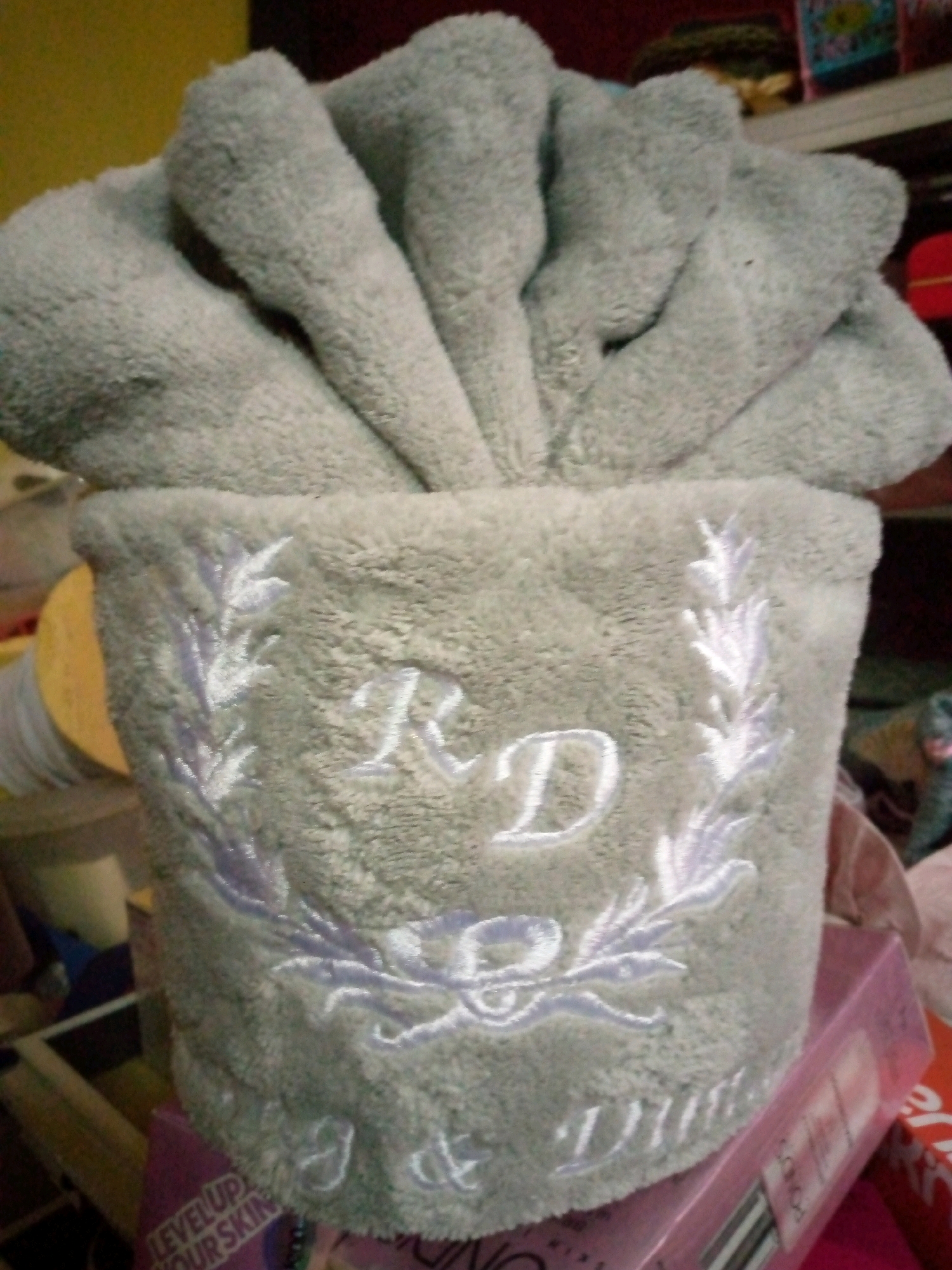 The image shows a close-up of a folded light gray towel arranged in a decorative manner, resembling a flower or fan. The towel is embroidered with a white and light purple laurel wreath design and the initials "RD". The towel is placed on top of a pink box with some text visible on its side, including the words "LEVEL UP YOUR SKIN". The background is out of focus, showing hints of shelves with colorful objects, suggesting an indoor setting, possibly a retail environment or a home storage area. The lighting appears to be from artificial sources, casting a warm glow on some surfaces. There are no people visible in the image. The location is not specified in the image or the provided context.