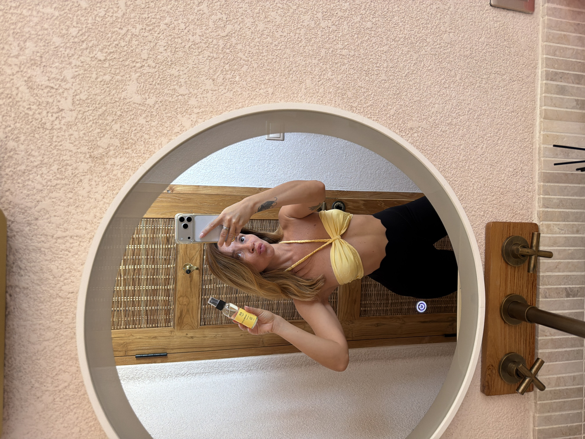 A woman takes a mirror selfie in what appears to be a bathroom. She is wearing a yellow bikini top and black leggings. In her left hand, she holds up a small bottle of what appears to be a skincare product. Her right hand holds her phone, which is angled to capture the photo. Reflected in the mirror behind her is a wooden door with a woven cane panel and a light switch. The bathroom walls have a textured, peachy-pink finish. Below the mirror, a wooden plank holds three brass-colored faucets. The location is not specified as being in Mengwi, Indonesia.