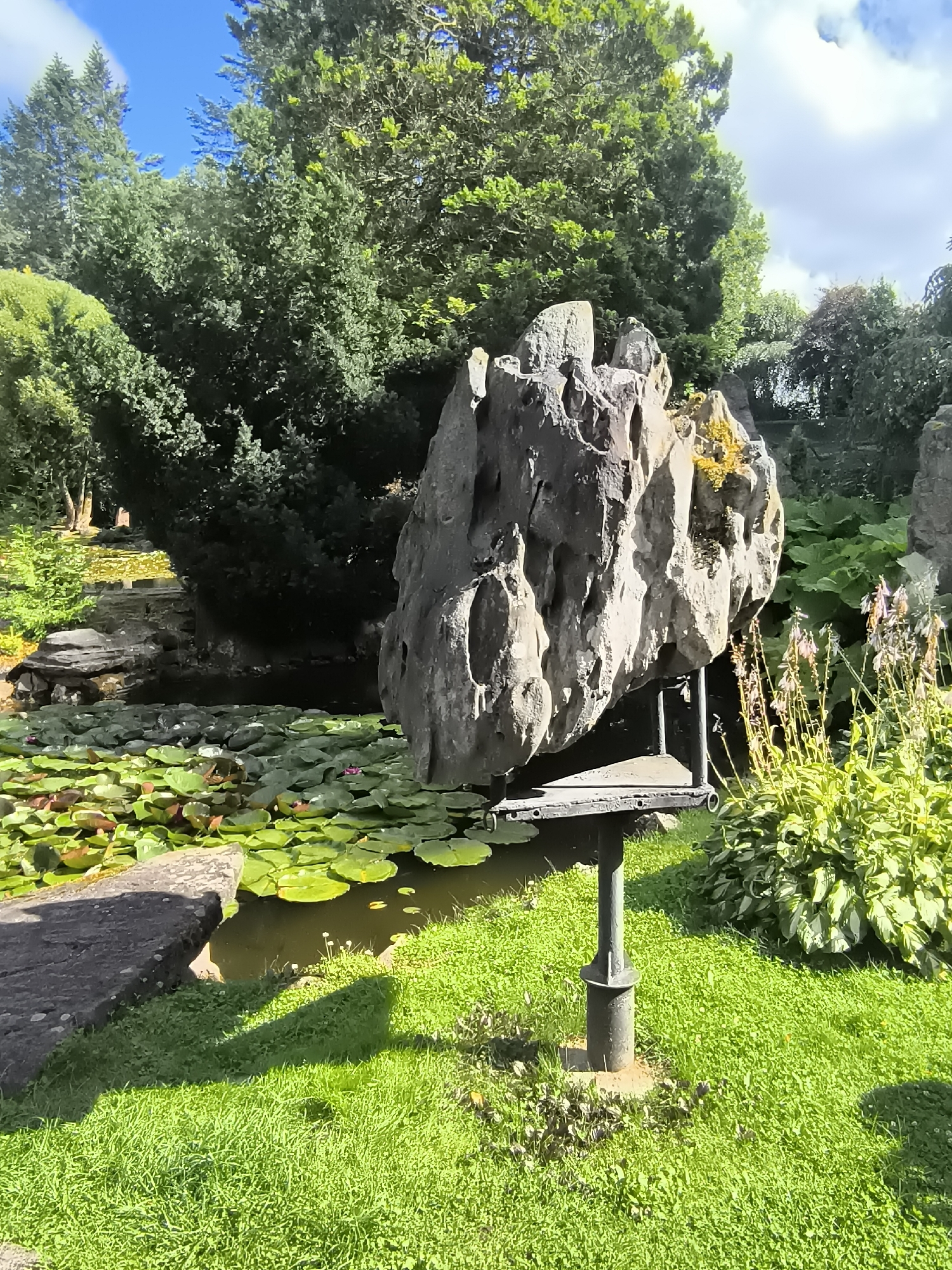 The image shows a garden with a large, uniquely shaped gray rock formation balanced on a metal stand in the foreground. To the left of the rock formation is a pond filled with numerous green lily pads and a few pink lily blossoms. A stone slab edge is visible in the lower left corner of the pond. Behind the pond and rock, lush green trees and foliage dominate the background, with a bright blue sky with scattered clouds visible above. The ground in front of the rock formation is covered in vibrant green grass. The scene appears to be a tranquil outdoor setting, possibly a park or botanical garden. This specific rock formation is a boulder from the Mosėdis boulder park in Lithuania.