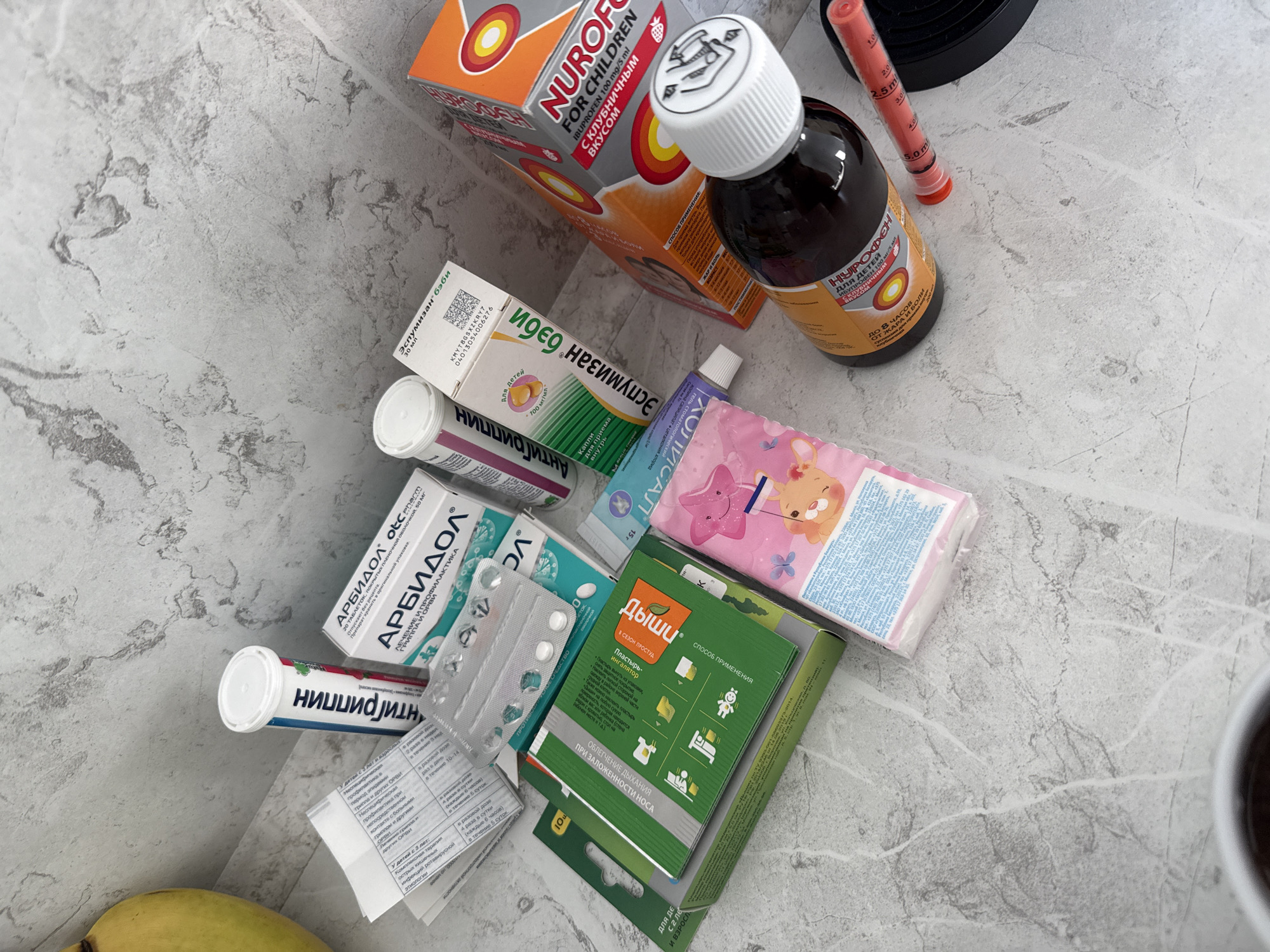 The image displays a collection of children's medications and related items laid out on a grey, speckled countertop, likely in a home environment. Visible items include:
* A white tube with blue and red lettering, identified as "АНТИГРИППИН" (Antigrippin), a medicine for flu and colds.
* A white box labeled "АРБИДОЛ" (Arbidol), a medicine used for treating and preventing influenza and other viral infections.
* A small white tube, possibly a medicated ointment or cream.
* A pink package with cartoon characters, likely containing wet wipes or tissues.
* Several blister packs with white pills.
* A green box with diagrams and text in Russian, appearing to be instructions or packaging for a medication.
* An orange box with red and yellow circles, labeled "NUROFEN FOR CHILDREN" with Cyrillic text indicating strawberry flavor.
* A dark brown glass bottle with an orange label, also identified as "NUROFEN" for children.
* A red plastic syringe-like dispenser with markings in milliliters, likely for liquid medication.
The scene does not provide any specific visual cues that would allow for identification of the location within Lyubertsy, Russia, or any details about people, actions, or interactions. It is a still life of medical supplies.