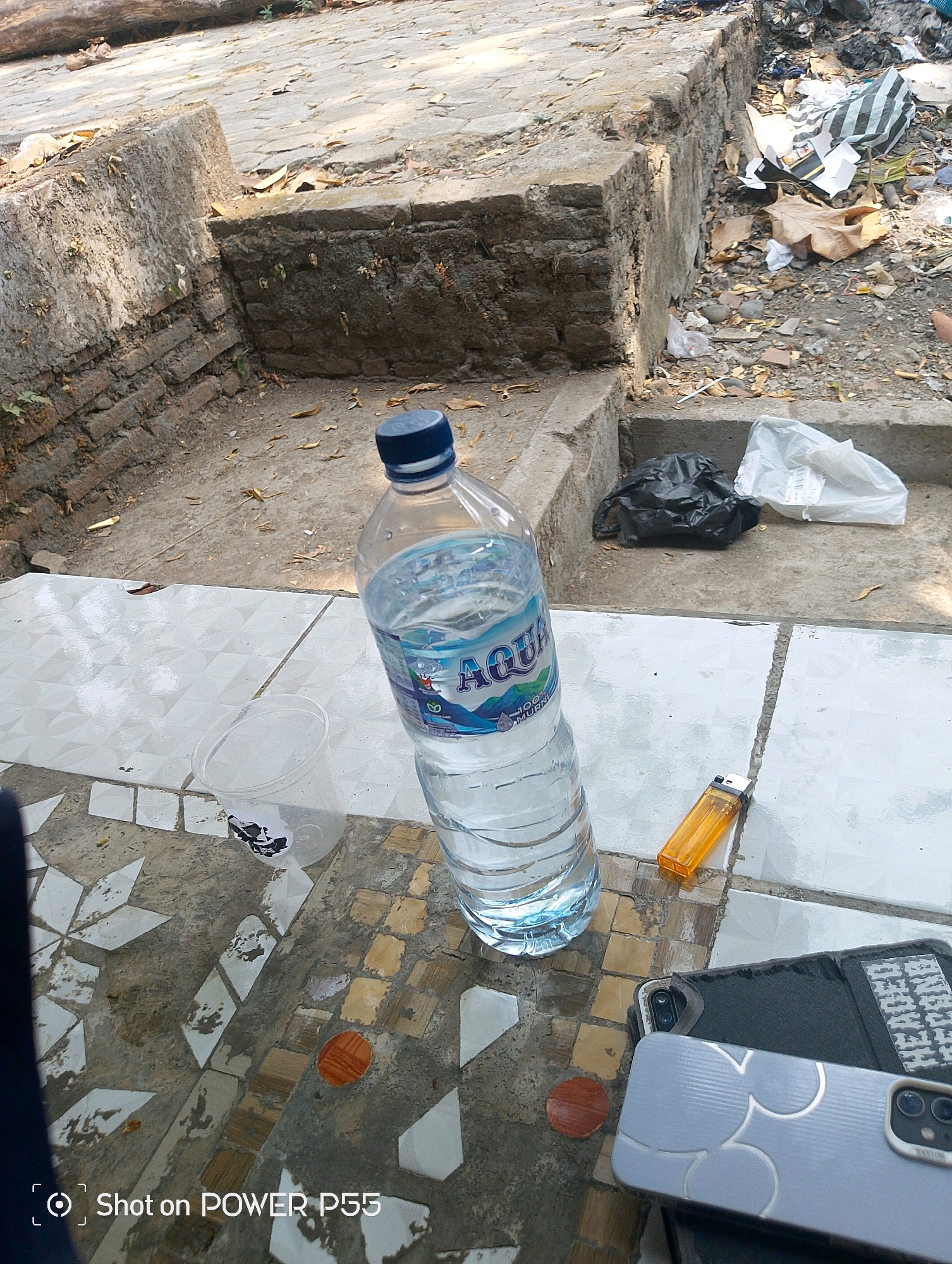 In Lebaksiu, Indonesia, a partially filled bottle of AQUA brand water stands upright on a tiled surface, its blue cap closed. Next to it rests an orange disposable lighter. In the background, a collection of trash bags, including a black one and a white one, are visible near a concrete structure. The ground is a mixture of concrete and stone, littered with dried leaves and debris, suggesting an outdoor urban environment. The scene appears to be captured from a slightly elevated perspective.