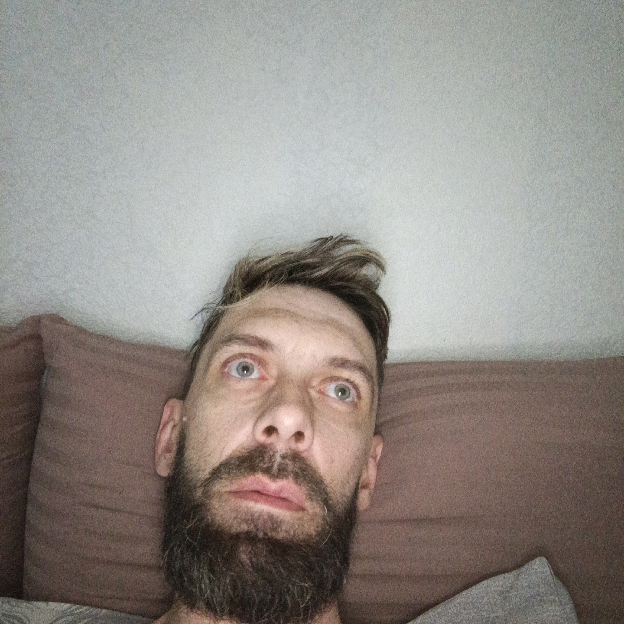 A man with a beard is lying down, looking upwards. He appears to be on a bed with a pillow. The background is a plain wall. No location information related to Марковский, Russia, is discernable.