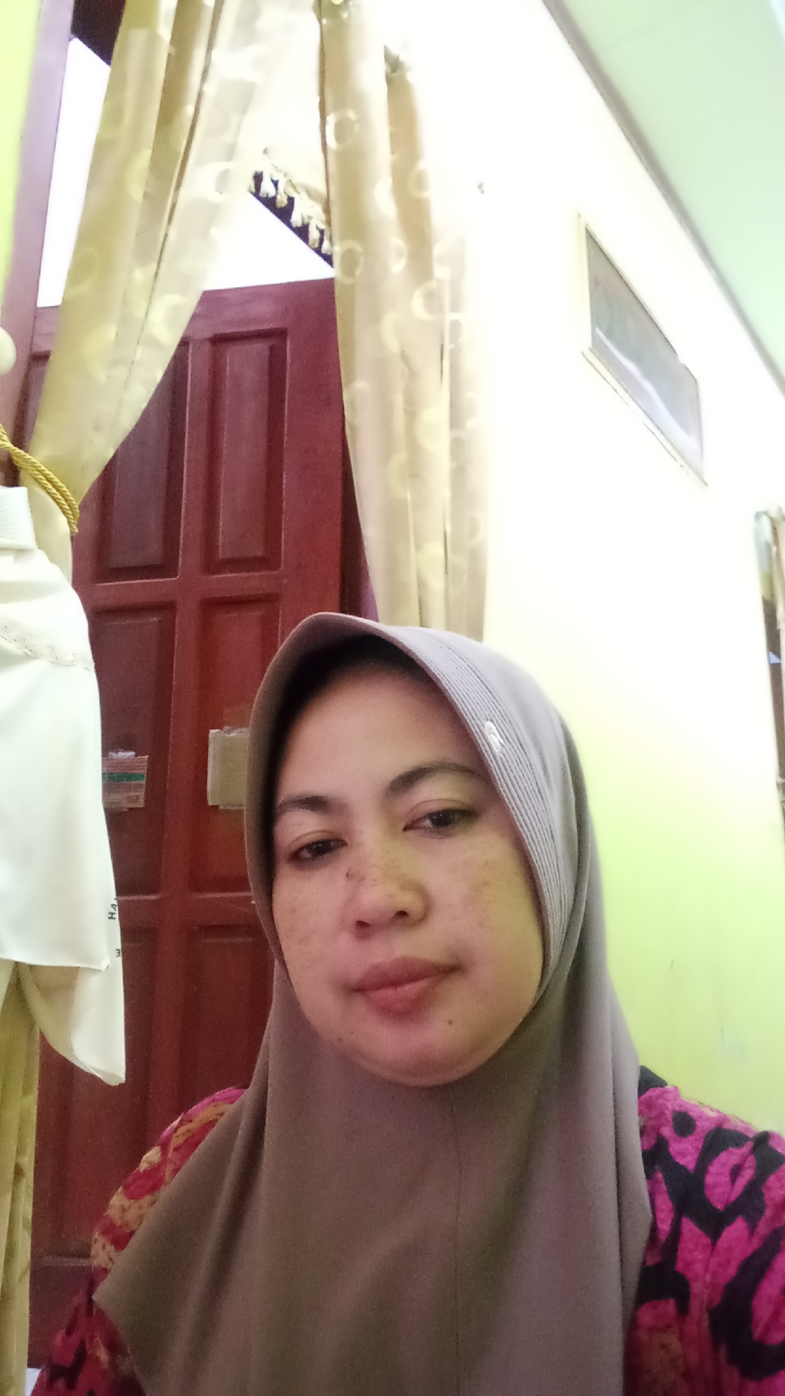 A woman wearing a grey hijab and a pink and black patterned shirt is taking a selfie in what appears to be a domestic setting. She is positioned in the foreground, with her face and upper body filling most of the frame. Behind her, a dark red wooden door with paneling is visible, partially obscured by light yellow curtains with circular patterns. To the right of the door and the woman, a light green wall is present. A window with a light-colored frame is mounted high on this wall, and a portion of the ceiling is also visible. The woman's expression is neutral, and she is looking directly at the camera. The lighting suggests an indoor environment, possibly during daylight hours. The image does not contain any explicit information about the city of Glenmore, Indonesia.