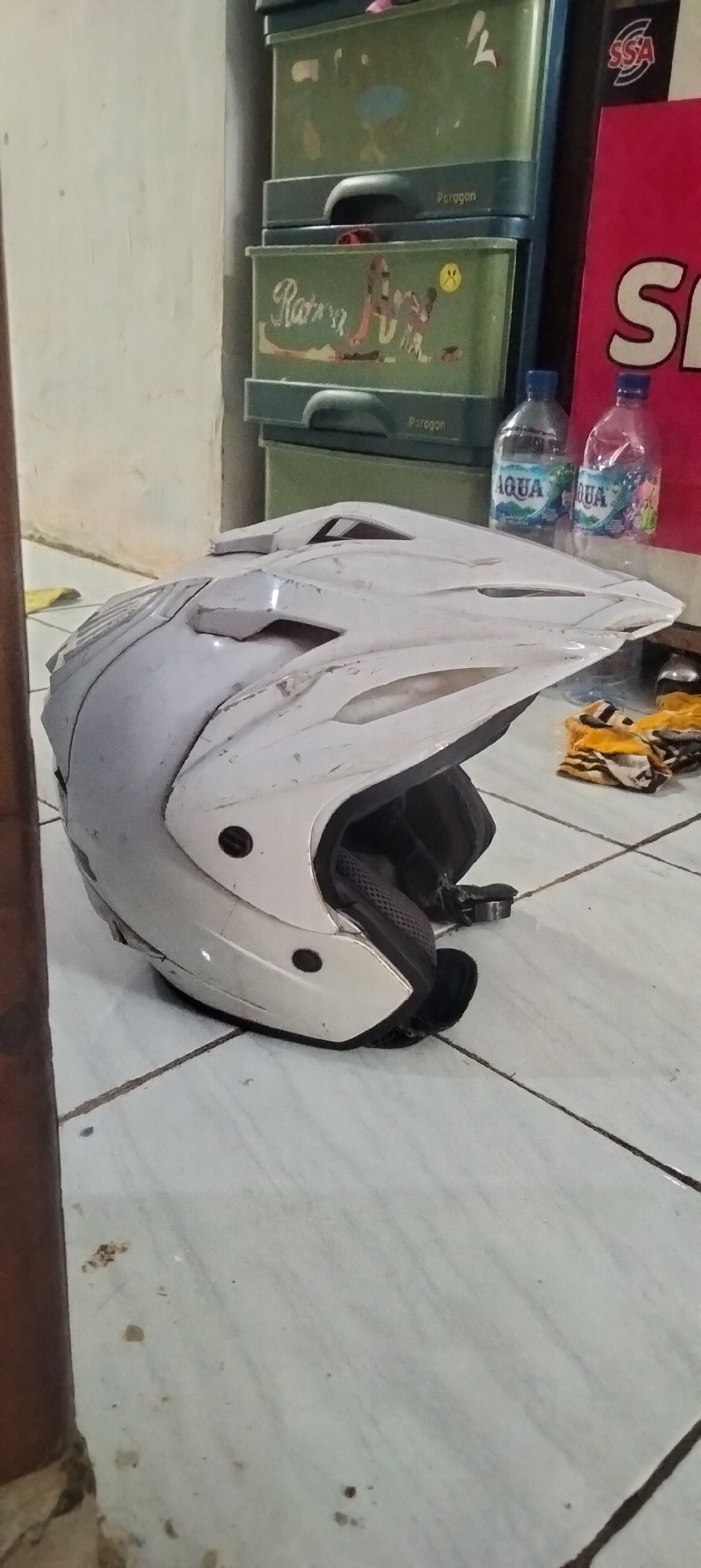 A white motocross helmet is positioned on a light-colored tiled floor. The helmet is slightly dusty and has visible scuff marks, indicating use. To the right of the helmet, there are two plastic bottles of Aqua brand bottled water. Behind the bottles and helmet, a stack of green plastic drawers is visible. The drawers have handles and some appear to have decorative stickers. The top drawer has "Paragon" printed on it, and a lower drawer features a drawing of a cat. Further to the right, a partially visible large red sign with white lettering that appears to say "S" is present. The overall environment suggests an indoor setting, possibly a storage area or a room in Antirogo, Indonesia.
