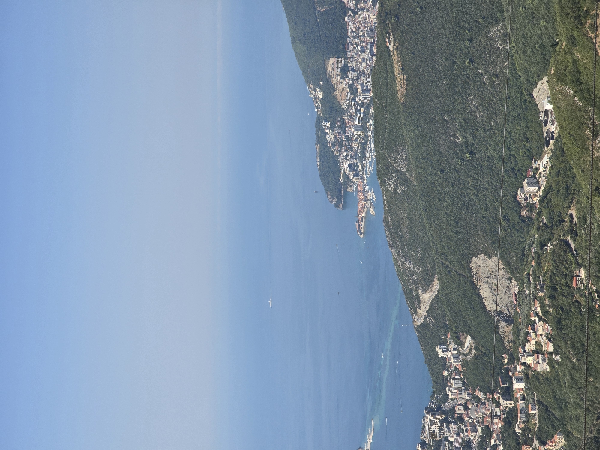 This is a high-angle, wide shot of a coastal city and the Adriatic Sea under a clear blue sky. The city, identified as Brajici, Montenegro, is nestled along the coastline and extends up the densely vegetated hillside. Buildings of varying sizes and colors, including residential and commercial structures, are visible throughout the urban area, with a prominent harbor filled with numerous boats. Roads wind their way through the city and up the steep slopes. The sea is a deep blue, with a few scattered boats and a larger vessel visible in the distance. The foreground is dominated by the lush green of the mountainous terrain, with some exposed rock faces. There are no people or animals readily apparent in the image. The overall impression is one of a scenic, developed coastal landscape.