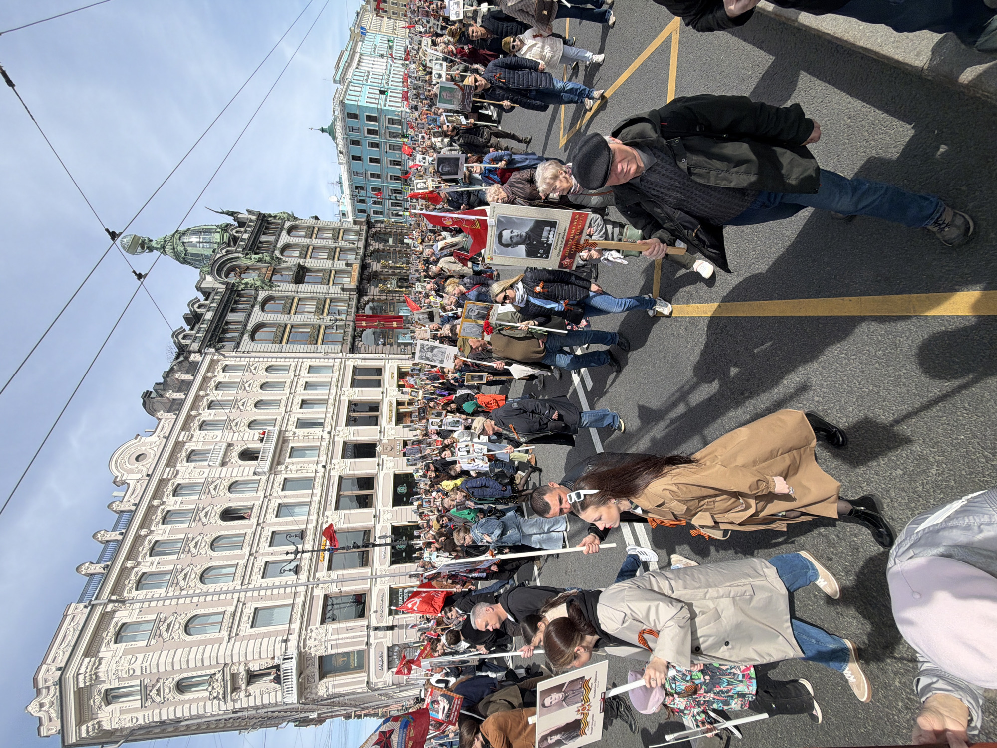 A large crowd of people is marching in a street procession in Saint Petersburg, Russia. The people are carrying portraits of individuals, some with military decorations, and red flags. They are walking along a street with yellow painted lines, with tall buildings on either side. The Singer House is visible in the background with its distinctive glass dome. Many people in the crowd have orange and black striped ribbons pinned to their clothing.