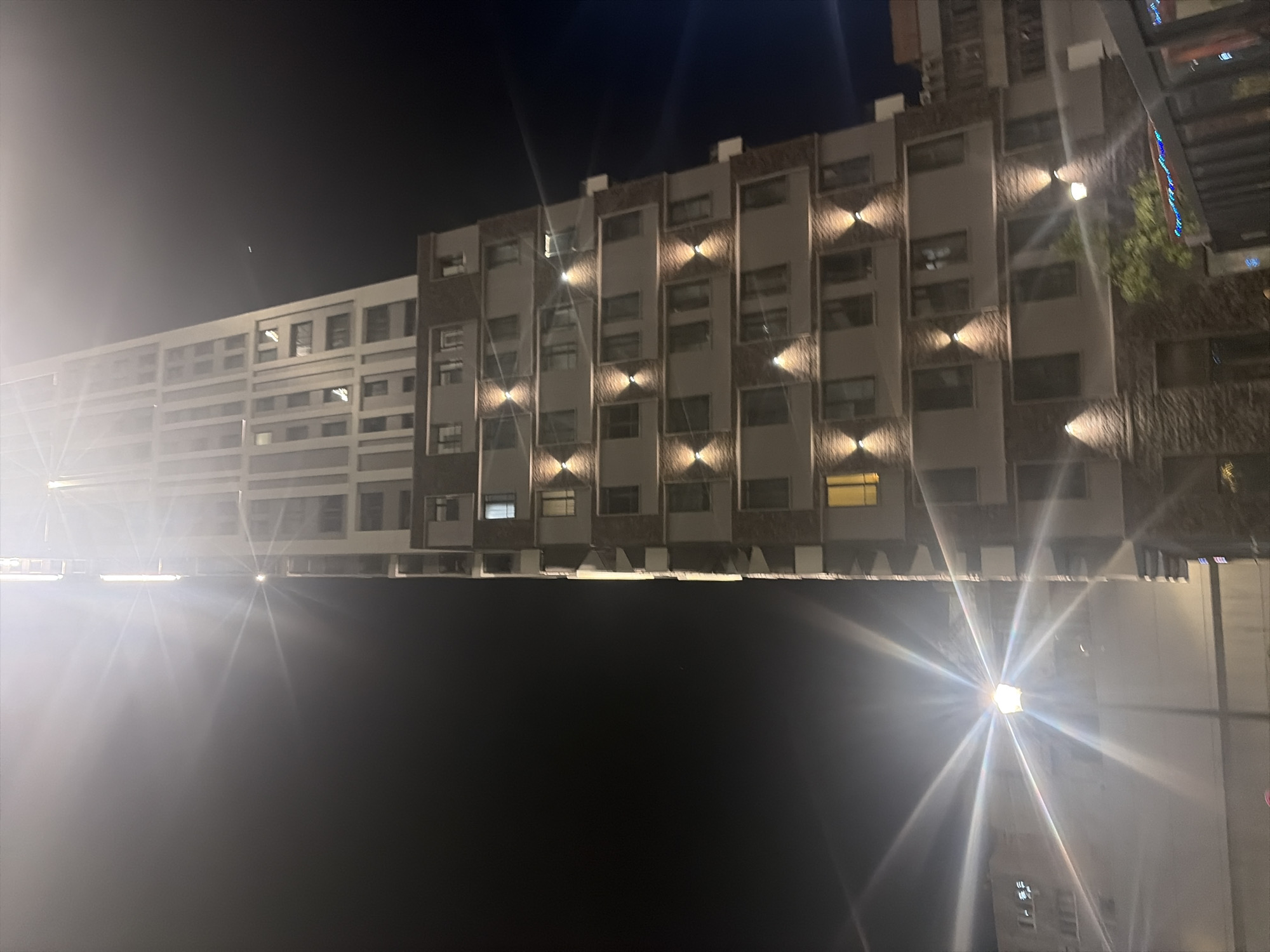An exterior low-angle vertical shot captures a modern high-rise apartment building at night in Moscow, Russia. The building is characterized by its light-colored facade with dark grey or brown brick accents and numerous windows. Exterior lighting illuminates various levels of the building, with strategically placed spotlights casting light upwards onto the facade. A prominent streetlamp with a hexagonal lantern and emanating light flares is visible in the foreground on the left side of the image. The background consists of a dark night sky, with the building's lights creating strong lens flares that dominate the upper portion of the image. Other buildings are partially visible to the left and right. The scene suggests a well-lit urban environment at night.