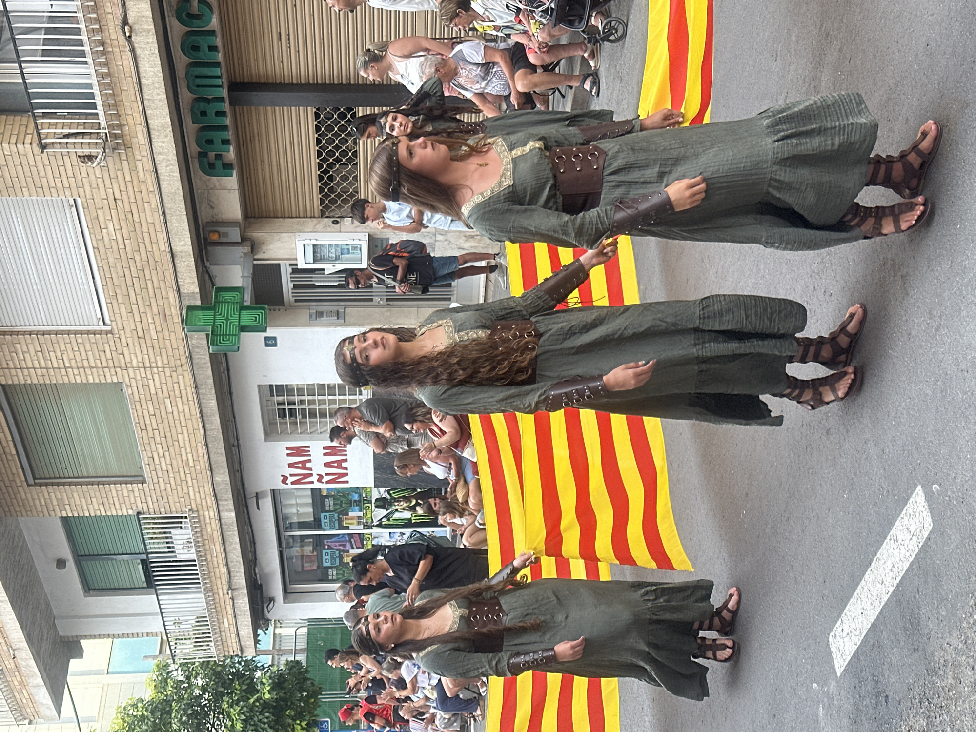 The image depicts a street scene in Alicante, Spain, likely during a festival or parade. Three young women, dressed in historically inspired green dresses with brown leather corsets and armbands, walk in procession. They are adorned with headbands. Behind them, a large banner with horizontal red and yellow stripes, resembling the Senyera (flag of Catalonia and Valencia), is unfurled.
In the background, a building with a brick facade houses shops. One prominent sign reads "FARMAC" with a green cross symbol, indicating a pharmacy. Another sign, partially visible, displays "ÑAM ÑAM" in red letters. Pedestrians are gathered on the sidewalk, some observing the procession, others engaged in conversation. The street is paved with asphalt and marked with a white line. The weather appears to be overcast.