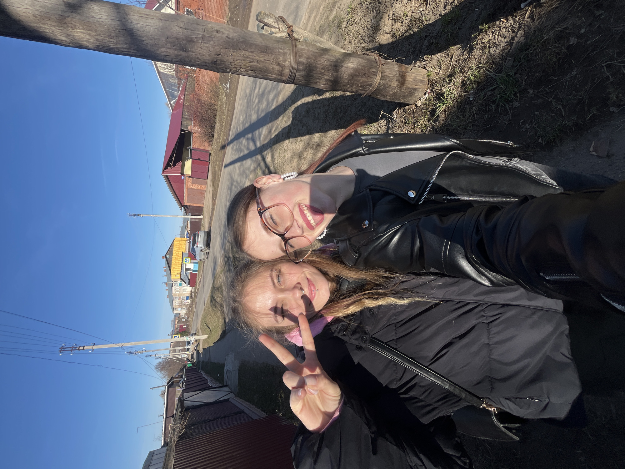 Two young women are captured in a bright, outdoor selfie, seemingly in the early spring or late autumn in Фоки, Russia. The image is oriented sideways, but the subjects are standing upright under clear, sunny skies.
The woman on the left, with dark hair, wears glasses and a black leather jacket over a grey top, adorned with a pearl necklace. She smiles broadly with her mouth open, showing her tongue. Next to her, on the right, is a woman with blonde hair, dressed in a black puffer jacket over a pink top. She flashes a peace sign with her right hand and has a nose piercing, also smiling. Both appear happy and relaxed, enjoying the sunny weather.
The background reveals a residential or rural street scene. Behind the women, a large wooden log or beam lies on the ground, casting a long shadow. Further back, various buildings with red roofs and brick walls are visible, along with bare trees and dry grassy patches. Utility poles with numerous power lines stretch across the frame, indicating an established infrastructure. A paved road or path runs alongside the buildings. The strong sunlight creates distinct shadows, suggesting midday or early afternoon. No discernible text is visible in the image.