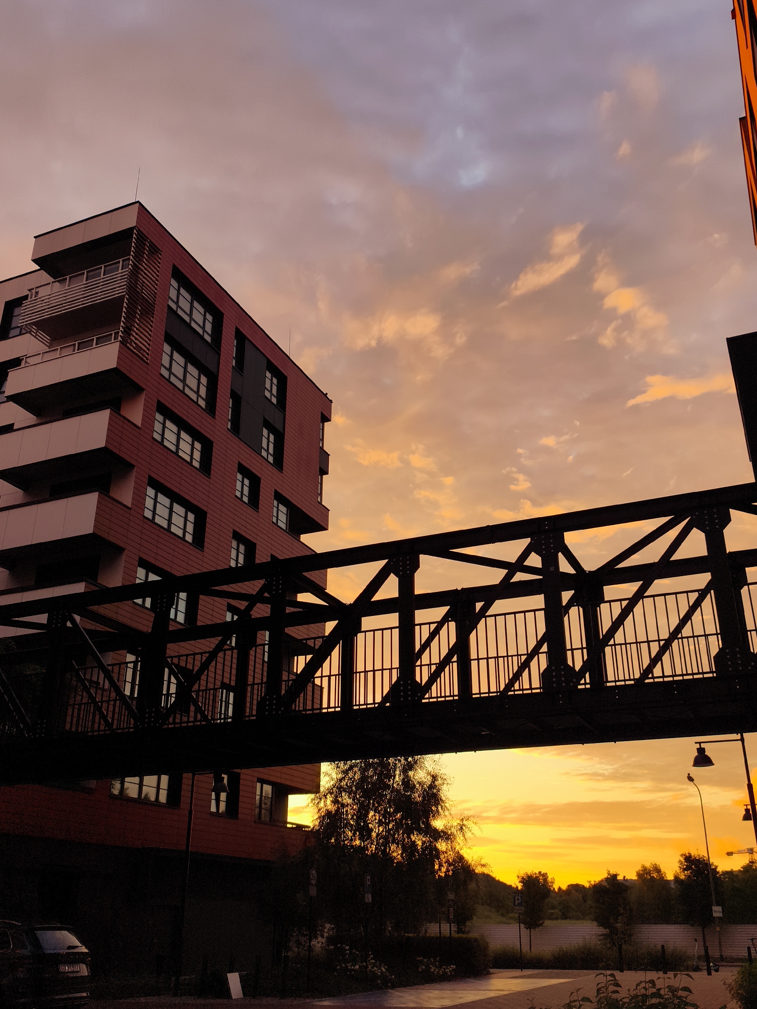 The image shows a modern apartment building in Gdańsk, Poland, bathed in the warm hues of a sunset. A dark, industrial-style pedestrian bridge stretches across the foreground, silhouetted against the colorful sky. The building, with its red and dark grey facade and balconies, is a prominent feature on the left side of the frame. The sky above is a canvas of orange, pink, and purple clouds, indicative of a beautiful sunset. The lower part of the image reveals a street-level view with trees, streetlights, and a car parked on the left. The overall scene captures an urban landscape at dusk, with a blend of contemporary architecture and the natural beauty of the sky.