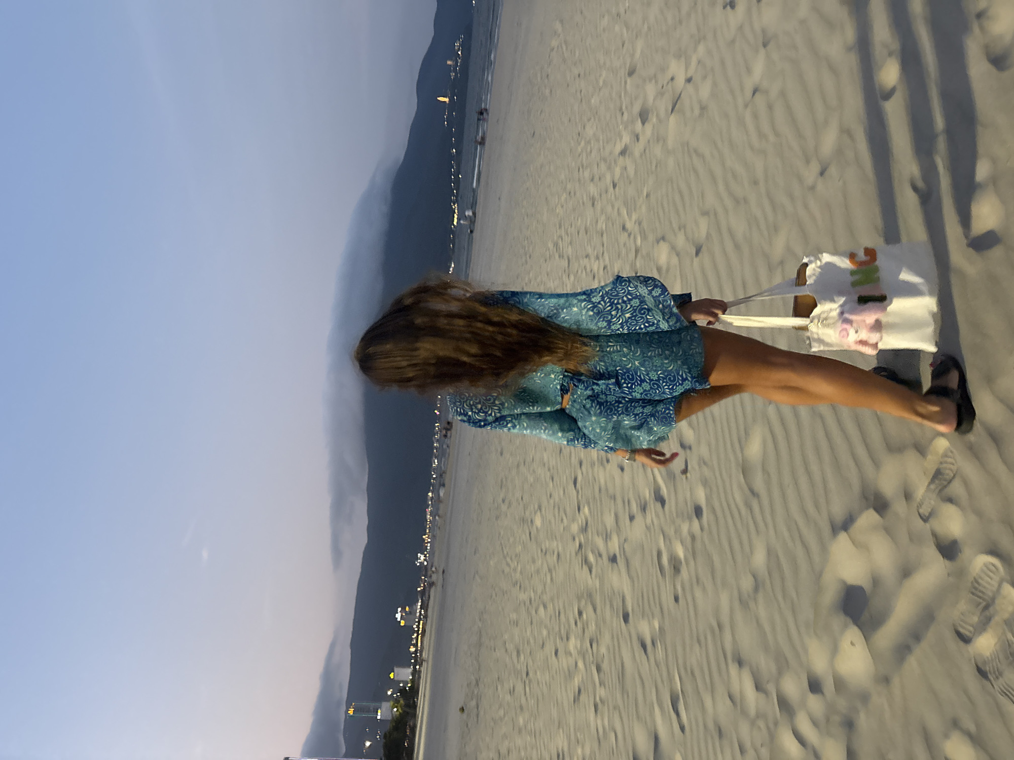 The image captures a person standing on a sandy beach in Da Nang, Vietnam, during dusk or early evening. The subject, viewed from behind, is a person with long, flowing brown hair, wearing a blue and white patterned cover-up or dress. They are carrying a white tote bag on their right side, which has visible text "I'M G" and a pinkish character graphic, possibly a pig or a similar stylized animal. Their feet are clad in dark sandals, and numerous footprints are visible in the sand.
The scene depicts a wide expanse of beach, with the ocean to the left of the person. In the distance, a prominent mountain range is silhouetted against the twilight sky, which shows gradients of light blue and grey. Along the base of the mountains and stretching along the coastline, a dense cluster of city lights indicates an urban area. The sky is clear, suggesting fair weather. The overall impression is one of tranquility at the end of the day, with the natural landscape blending with the distant cityscape.