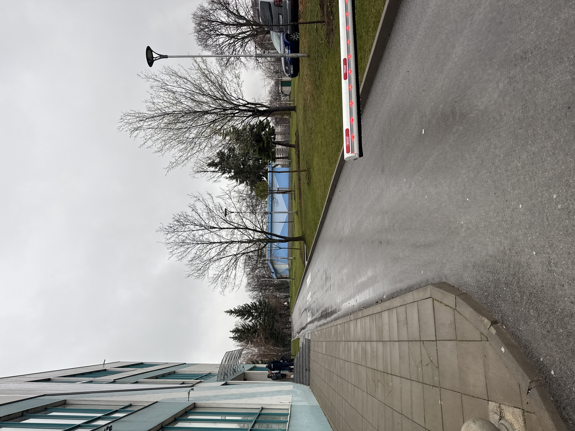 This image depicts an outdoor scene in Sofia, Bulgaria, under an overcast sky, with visibly wet asphalt and paved surfaces. On the left, a multi-story building features a facade with alternating light blue and white horizontal stripes, punctuated by vertical sections of windows with teal frames. A set of concrete steps leads to an entrance covered by a transparent, curved awning. Approximately five individuals are observed on these steps and at the entrance; one individual wears a dark jacket displaying white "POLICE" text on the back. A gray tiled sidewalk runs alongside the building, bordering a dark, wet asphalt road that appears to curve uphill. A white traffic barrier arm, marked with "ROGER" text and equipped with multiple illuminated red LEDs, extends horizontally across the right side of the road, indicating a closed state. Beyond the road, a landscaped area of green grass contains several bare deciduous trees and a cluster of evergreen conifers. A dark, modern-style street lamp is positioned among the trees. In the middle ground, two vehicles are parked on the grass: a blue sedan and a silver minivan. Two cylindrical structures, painted with blue and white stripes, are visible in the distant background within the grassy expanse.