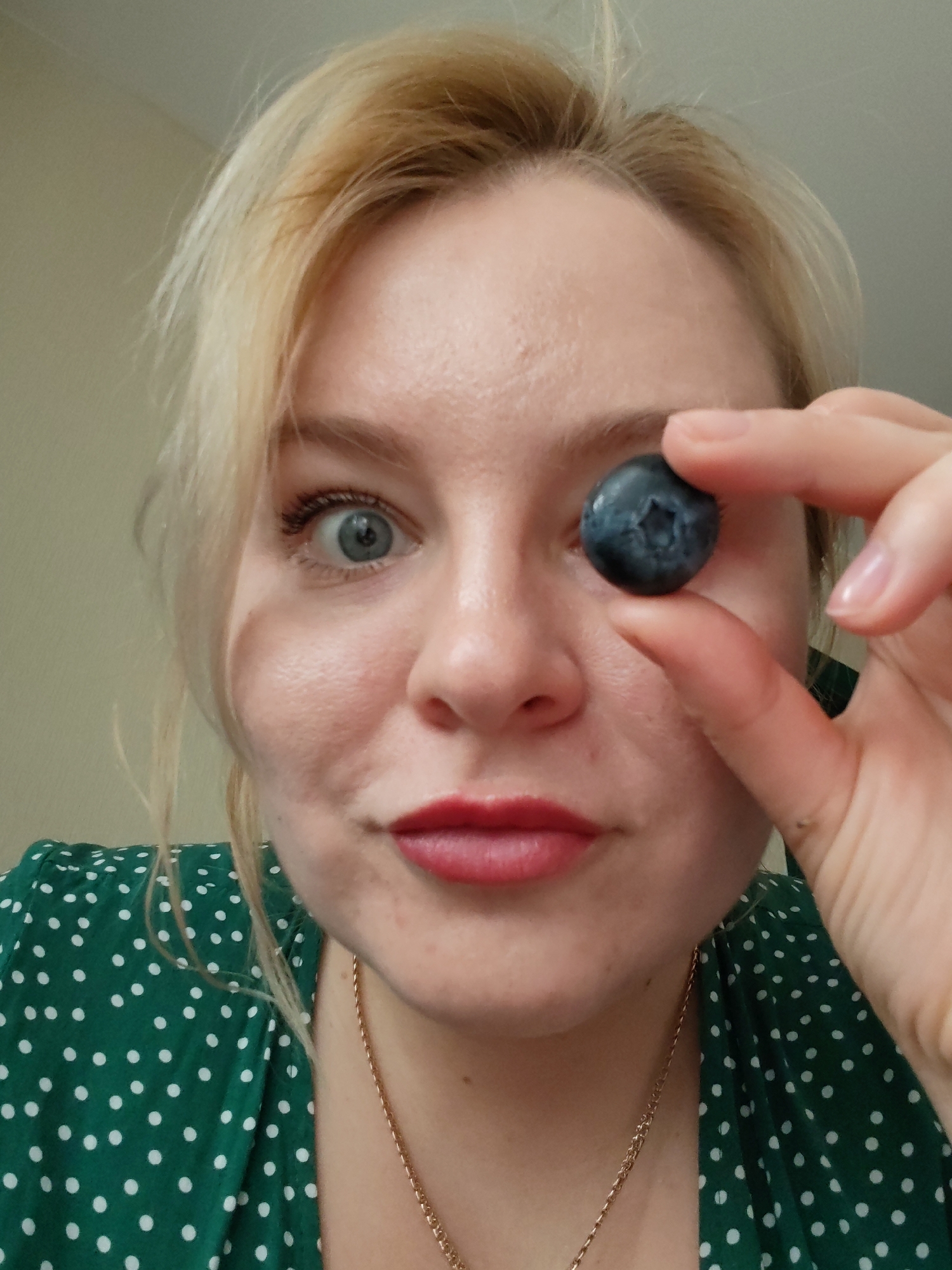 A close-up, eye-level indoor shot features a light-skinned woman with blonde hair looking directly at the camera. She holds a single dark blue blueberry between her thumb and index finger, positioned directly in front of her right eye, playfully obscuring it. Her left eye is wide open, gazing intently at the viewer, suggesting surprise or playful engagement. Her lips are painted with red lipstick and slightly pursed. She is wearing a green dress with white polka dots and a thin gold chain necklace. The background consists of a plain, light-colored wall, indicating an indoor setting. The lighting is soft and even, typical of an interior space. The overall mood of the image is lighthearted and quirky. There is no visible text in the image.