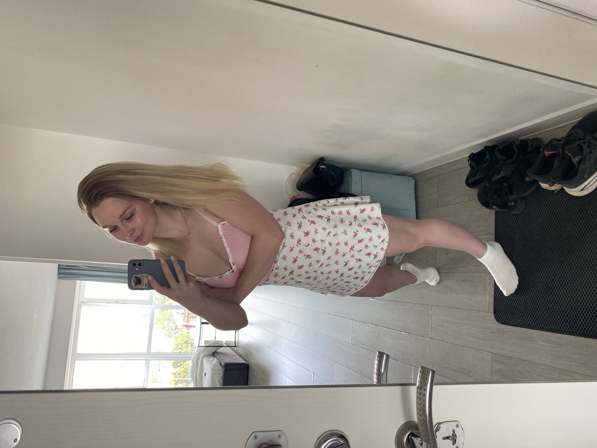 A young woman with blonde hair is taking a mirror selfie in what appears to be a bedroom or hallway. She is wearing a pink camisole with white lace trim and a white skirt with a floral pattern. She is also wearing white socks. In the background, through a large window, city buildings and trees are visible. There is a bed with white linens in the room. In the foreground, a black doormat is visible, with several pairs of shoes placed on it and beside it. The woman is holding a smartphone in her right hand, which has a phone case with a pug design. She is looking at her reflection in the mirror. The flooring is light-colored wood laminate. There are door handles visible on a white door on the left side of the image. A light blue storage box is located to the right of the woman. The scene does not provide any specific visual cues to identify its location within the city of Izhevsk, Russia.