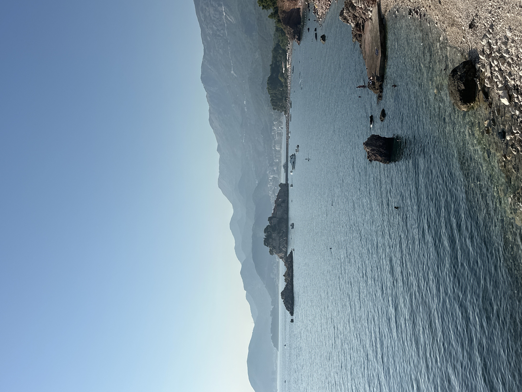The image depicts a serene coastal scene in Sveti Stefan, Montenegro. The foreground features a rocky shoreline with clear, blue water lapping against it. Several large, dark rocks are scattered in the shallow water, some of which are partially submerged. A concrete platform juts out into the sea on the right side of the image, with a few individuals visible on it and in the water.
Further out in the bay, the water is a deeper blue and appears calm. Small boats, including what looks like a yacht, are dotted across the water. In the middle ground, a cluster of rocky islets rises from the sea, with a small settlement visible on the largest islet, characterized by traditional buildings. The background is dominated by rolling, green mountains that fade into a hazy blue sky. The overall atmosphere is tranquil and picturesque, indicative of a warm, sunny day at the beach.