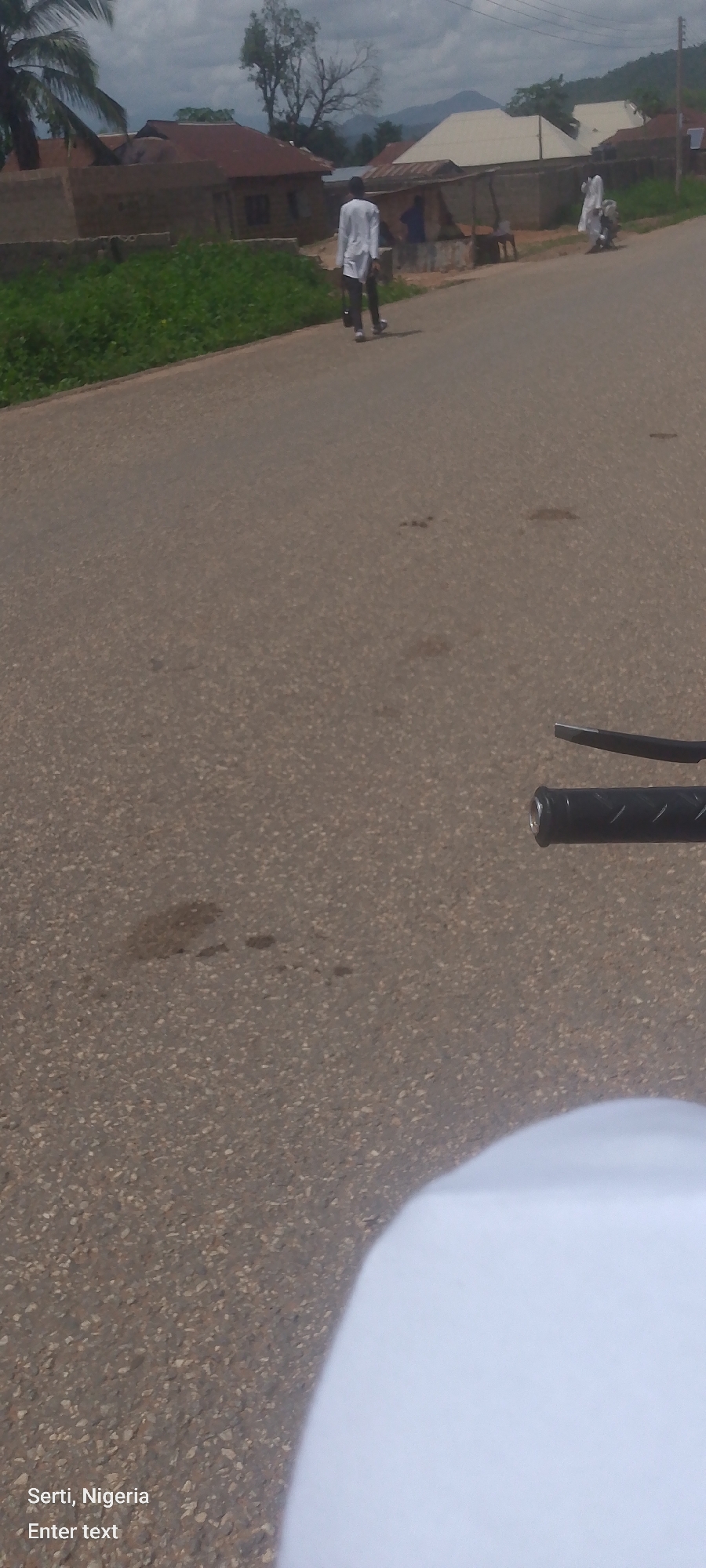 The image captures a scene on a paved road in Sertı, Nigeria. The road surface is asphalt with scattered dark patches. In the background, there are buildings with corrugated iron roofs, some appearing to be houses or shops. Vegetation, including trees and bushes, lines the side of the road. The sky is cloudy.
Several people are visible. One person, dressed in white traditional attire and carrying a bag, walks away from the camera. Further down the road, another individual in similar white clothing is on a motorcycle, possibly a passenger. The foreground shows a portion of a motorcycle's handlebars, suggesting the photo was taken from a moving motorcycle. The text "Serti, Nigeria Enter text" is overlaid on the lower left portion of the image.