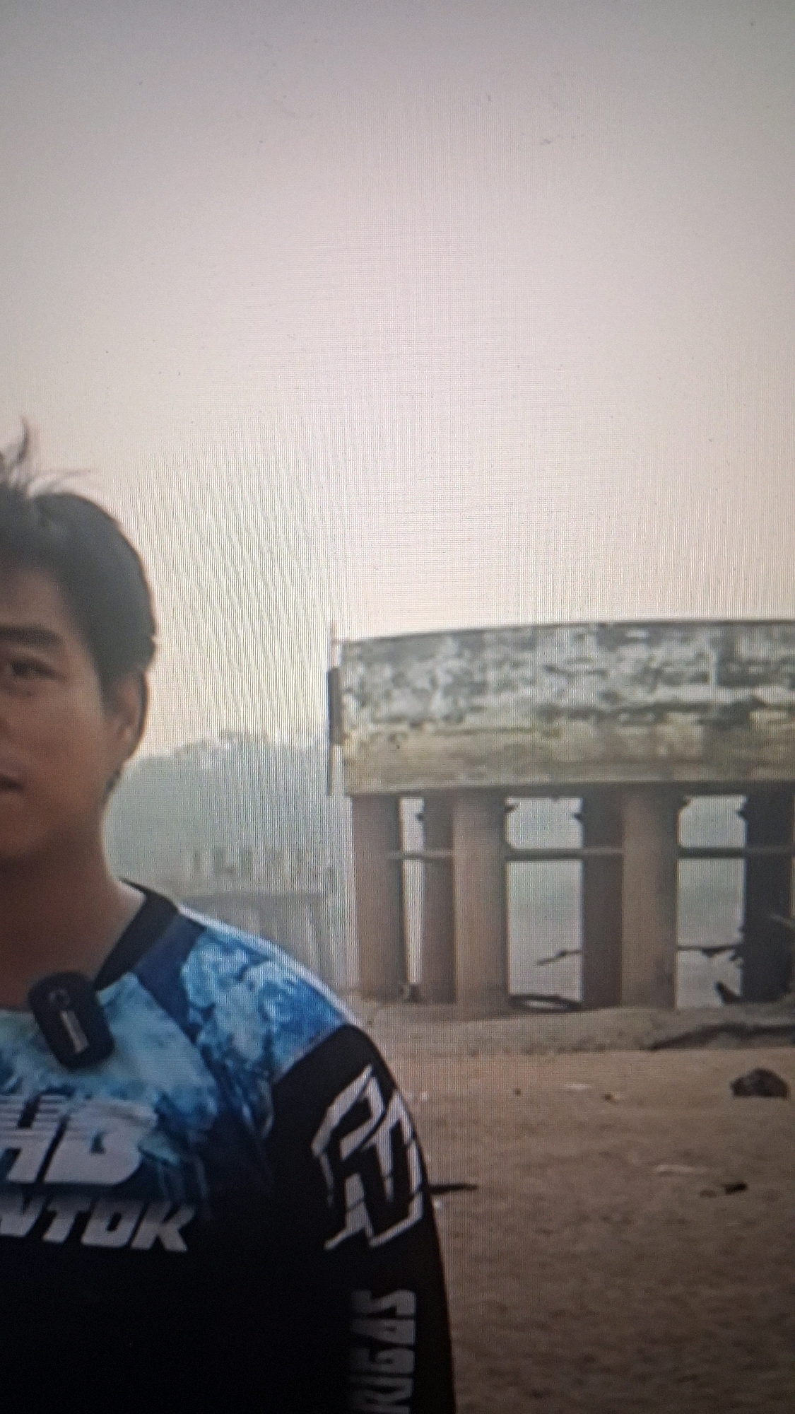A man is standing on a sandy area, possibly a beach or riverbank, with a large, weathered concrete structure in the background. The structure appears to be supported by thick, cylindrical pillars and has a flat, rectangular top. The man is wearing a dark blue and black long-sleeved shirt with a tie-dye pattern on the shoulders and sleeves, featuring white text and graphics. A small black device, possibly a microphone or camera, is clipped to his shirt collar. The sky is overcast and hazy, suggesting a diffused light. The overall scene appears to be outdoors, with natural elements like sand and possibly vegetation visible in the distance. The location is in the city of Bandung, Indonesia.