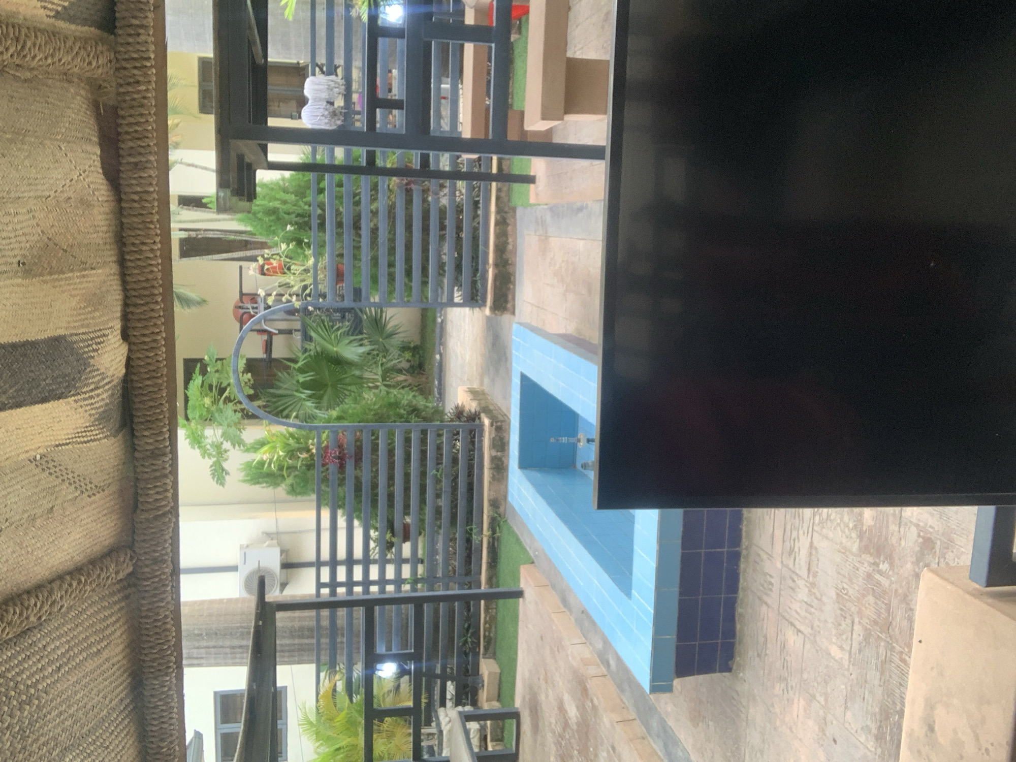 This media file captures an outdoor scene, likely in Abuja, Nigeria, featuring a small, tiled swimming pool with blue tiles. The pool appears empty and is situated on a paved patio area. Adjacent to the pool is a dark, solid structure, possibly a wall or a large piece of furniture, that dominates the right side of the frame.
To the left of the pool, a metal fence or railing with horizontal bars runs across the image, partially obscuring a garden area with lush green plants and palm trees. Beyond the greenery, a building with light-colored walls and windows is visible. An outdoor air conditioning unit is mounted on one of the walls.
In the foreground, on the far left, a woven textured material, possibly part of a thatched roof or a large basket, takes up a significant portion of the frame, providing a sense of an outdoor living or seating area. The lighting suggests it is daytime, with no strong shadows indicating a specific time. The overall impression is of a relaxed, perhaps residential or resort-like, outdoor space. There is no visible text in the image.