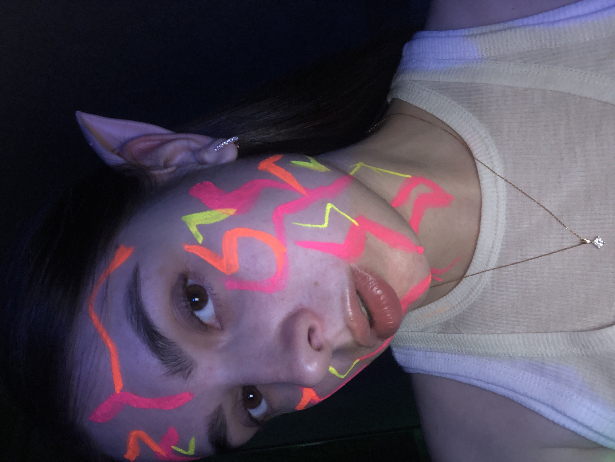A tilted portrait image captures a person in a low-light setting, possibly in Noviy, Russia, adorned with vibrant neon face and neck paint. The subject, likely female, has long dark hair and is wearing a light-colored, ribbed tank top or camisole. Their face and upper body are covered in glowing, abstract patterns of neon pink, orange, and yellow paint, which illuminate intensely under what appears to be blacklight.
The person features notable pointed, prosthetic ears, suggesting an elf or fantasy-themed costume. A delicate gold chain necklace with a small, clear stone pendant is visible around their neck. One hand shows a ring with small, clear stones. The subject looks directly at the camera with a neutral expression, their lips appearing glossy. The strong blacklight effect gives the skin a slightly purplish hue and makes the paint patterns pop against the dark background. The scene suggests a themed event such as a party, club, or rave, where blacklight and costume elements are prominent. There is no visible text in the image.