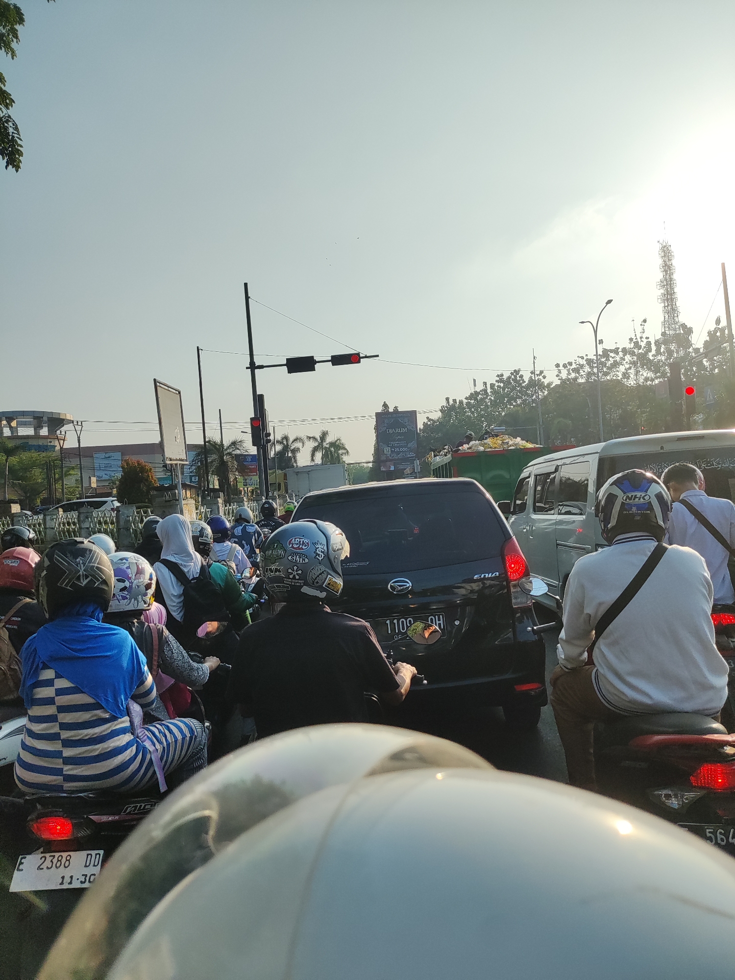 A street scene in Cirebon, Indonesia, captured on a bright day with a clear, pale sky. The image depicts a traffic jam with numerous motorcycles and a few cars. People on motorcycles, most wearing helmets, are stationary in the traffic. The foreground is dominated by the back of a white motorcycle helmet. In the middle ground, a black SUV is visible, followed by a silver minivan. A green garbage truck is parked on the right. Behind the traffic, buildings, palm trees, and a tall telecommunications tower are seen. A traffic light with a red signal is also present. The overall impression is a busy urban environment experiencing a traffic standstill.