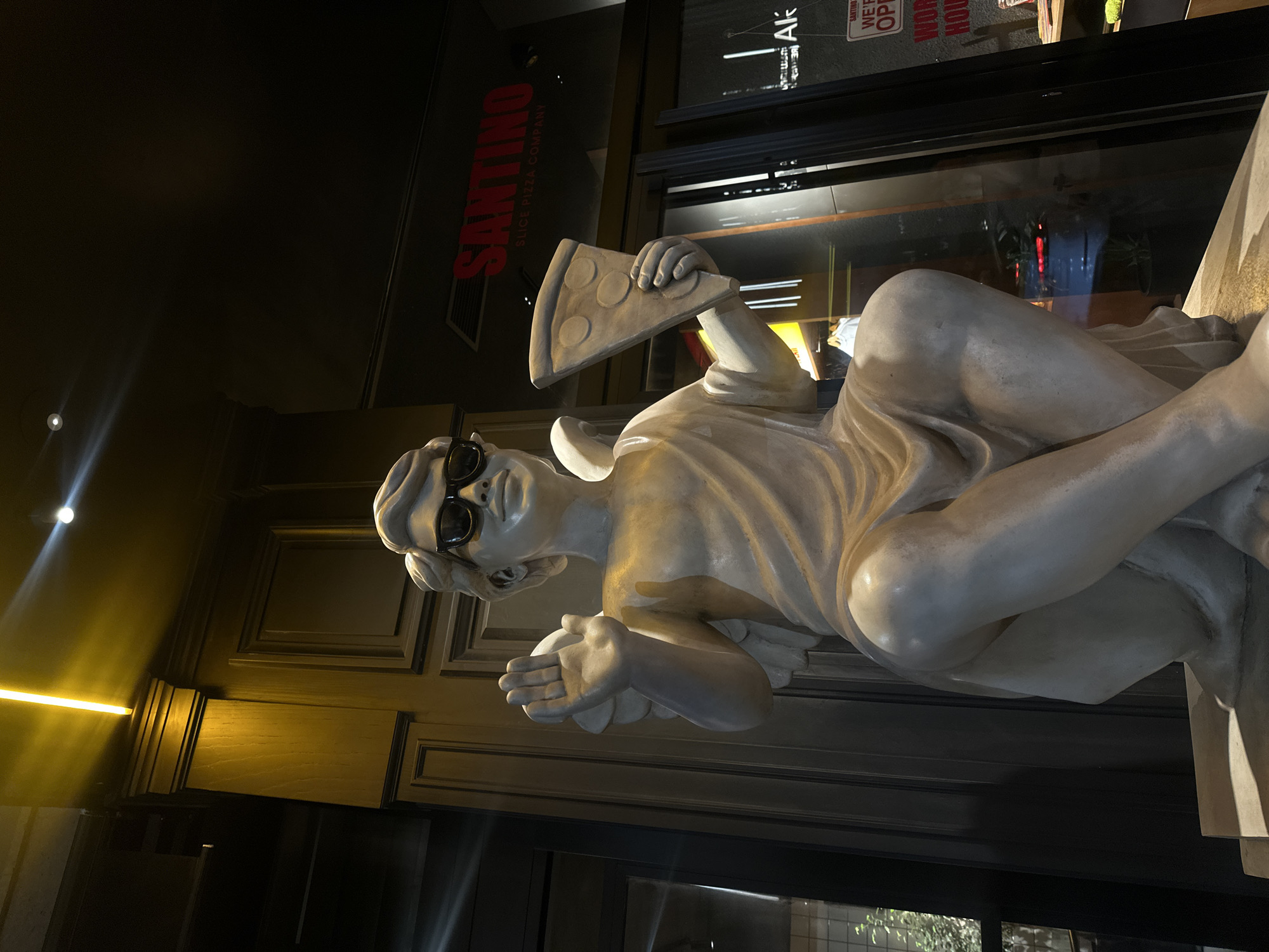 A sideways-oriented, long shot shows a statue of a Greco-Roman figure wearing sunglasses and holding a slice of pizza, positioned outside of Santino Slice Pizza Company in Yerevan, Armenia. The statue is made of light gray material. It reclines on a stone-like ledge. The pizza slice is held in the figure's right hand. The restaurant's sign, "SANTINO SLICE PIZZA COMPANY", is visible in red behind the statue. A door/window with signs indicating "WE'RE OPEN" and "WORK HOURS" can be seen in the background. The light is dim, suggesting it is nighttime.