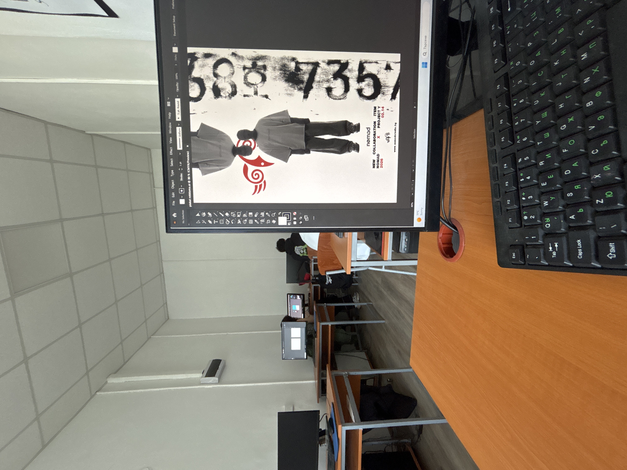 This image captures a brightly lit computer lab or classroom in Troyan, Bulgaria, with multiple students engaged in computer-based work. The photograph is taken from a slight aerial perspective, looking down on the workstations.
In the foreground, a black computer monitor displays a graphic design project, likely within Adobe Illustrator, given the "plakat rekklama.ai" filename visible in the software's interface. The design features a light background with large, distressed black numbers "585735" at the top. Below these numbers are two stylized figures dressed in grey garments, facing each other, with a red, organic-shaped symbol beneath them. Various text elements are incorporated into the design, including "nomad", "NEW COLLABORATION", "NOMAD X PROJECT Y", "2026", "ITEM", "02.14", and the website "www.aomadstudio.bg", which confirms a Bulgarian connection. The monitor's taskbar also shows local weather information: "11°C Sunny".
To the right of the monitor, a black keyboard with both Latin and Cyrillic characters is visible on a light brown wooden desk. A small red cylindrical holder is embedded in the desk near the keyboard. In the mid-ground and background, other students are seen working at similar computer setups, with their monitors displaying various interfaces. One black Reebok backpack is noticeable on a chair in the background. The room features grey walls, a white ceiling with square acoustic panels, and multiple desks arranged in rows. A rectangular, metallic speaker or ventilation unit is mounted on the wall.
