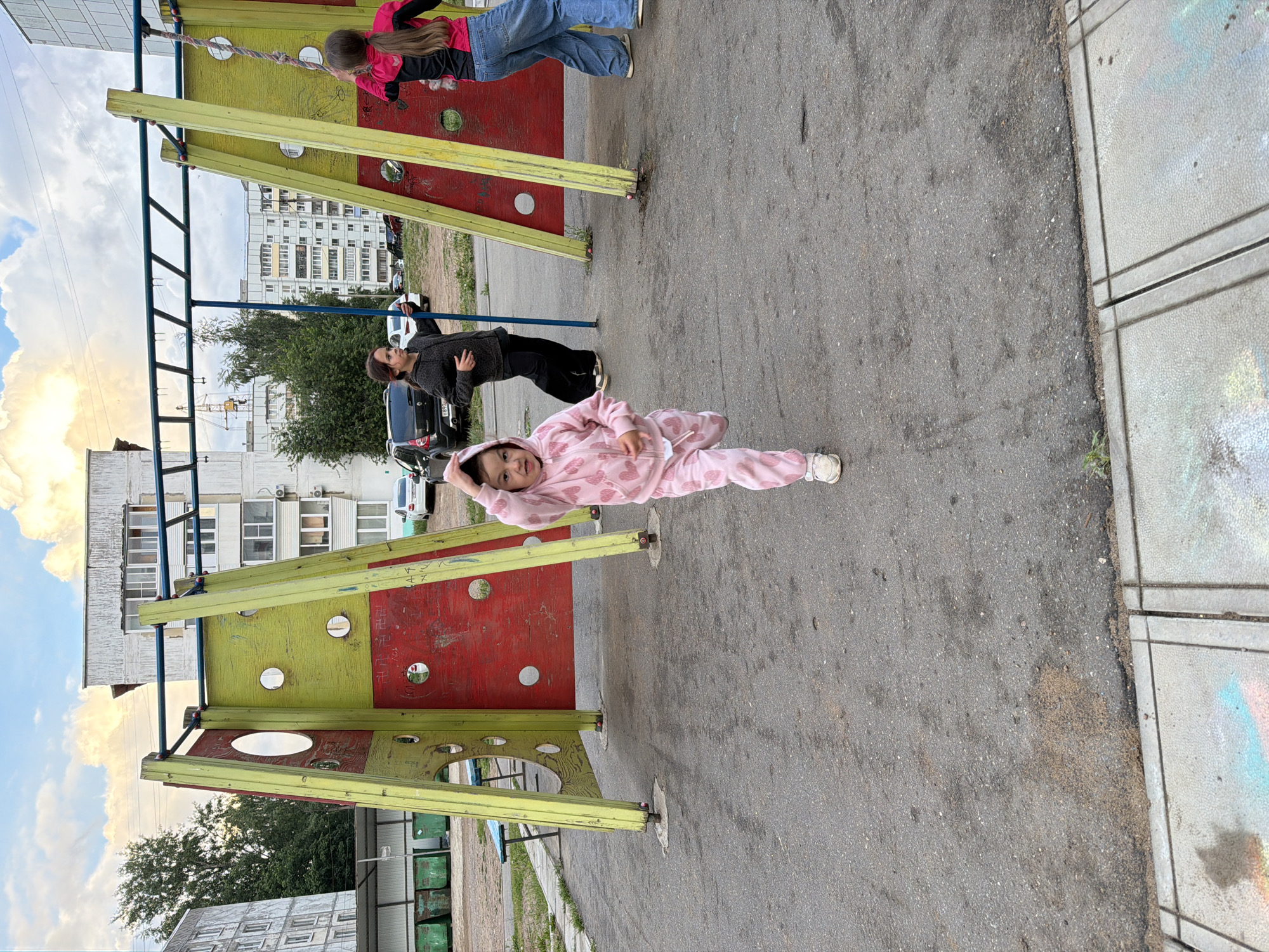 This portrait-oriented image, rotated 90 degrees counter-clockwise, depicts an outdoor playground scene in what appears to be an urban residential area of Chaikovsky, Russia.
In the foreground, a young child wearing a pink jumpsuit with white heart patterns is lying on an asphalt surface, smiling and looking directly at the camera. Her hands are near her head, and her white shoes are visible. To her right, a concrete sidewalk is partially visible.
Behind the child is a colorful playground structure with yellow frames and red panels. Another person, a girl or young woman in a pink top and blue jeans, is actively climbing or standing on the upper part of this structure, facing away from the camera. Further back and to the left of the climbing structure, a third person in dark clothing (dark top and pants) stands on the asphalt, looking towards the left.
The background features multi-story apartment buildings, characteristic of a Russian cityscape, with some trees providing greenery. The sky is partly cloudy, indicating daytime with diffused light. On the red and yellow panels of the playground equipment, some light-colored graffiti or markings, including what appears to be Cyrillic text, are visible. The scene captures a moment of play and relaxation in a community setting.