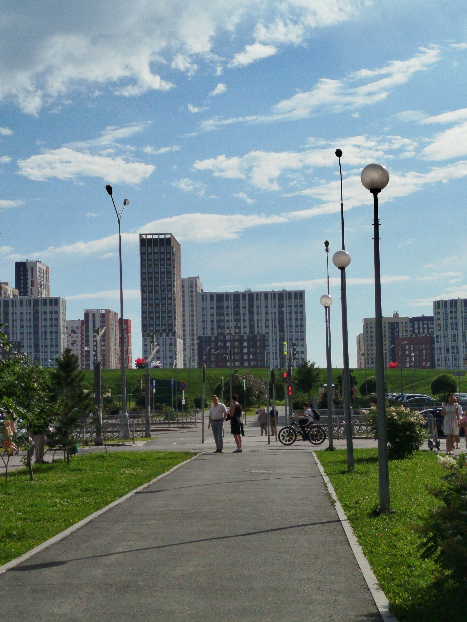 In Yekaterinburg, Russia, a wide, paved walkway runs diagonally through a well-maintained park area with lush green grass and scattered trees. The sky above is a bright blue, dotted with fluffy white clouds. Tall, modern apartment buildings, characteristic of urban development, line the background.
On the walkway, several people are present. A man in a light-colored shirt and dark pants walks towards the camera, while a woman in a dark dress walks away from him. Further down the path, a person on a bicycle is visible. A few other individuals are scattered throughout the scene.
Streetlights, some with distinctive spherical globes, stand along the walkway, indicating the area is equipped for evening use. Beyond the walkway and grass, a road with parked cars is visible, suggesting the park is adjacent to a residential or commercial area. The overall impression is of a clean, modern urban environment designed for recreation and living.