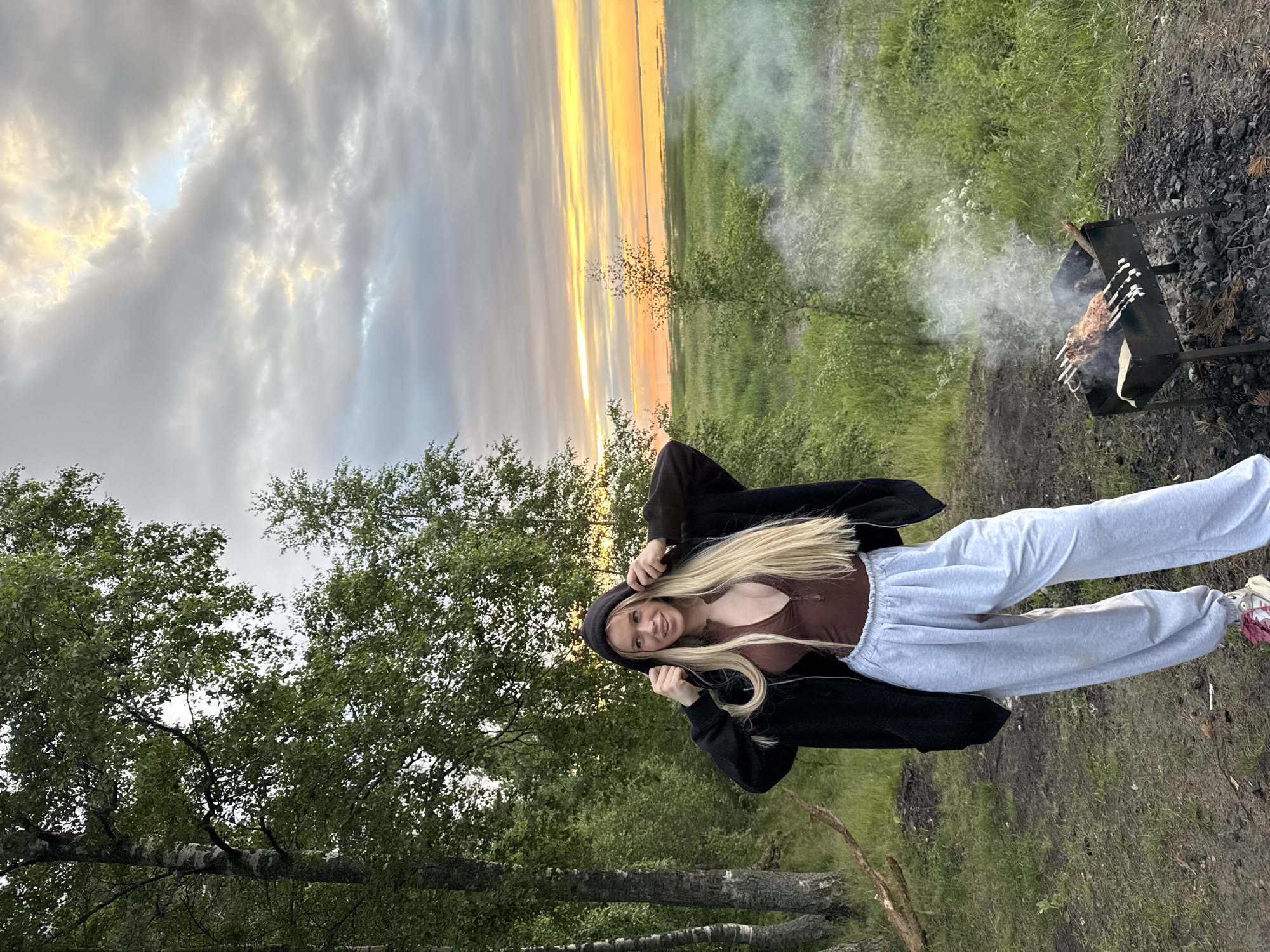 A young woman with long blonde hair stands in a clearing near trees, wearing a dark hooded jacket, a brown top, and light grey sweatpants. She is holding the hood of her jacket with both hands. In the foreground to her right, a metal grill with meat skewers is smoking. The background features a grassy field and a body of water under a cloudy sky with sunset colors. This scene is located in Bolshaya Izhora, Russia.