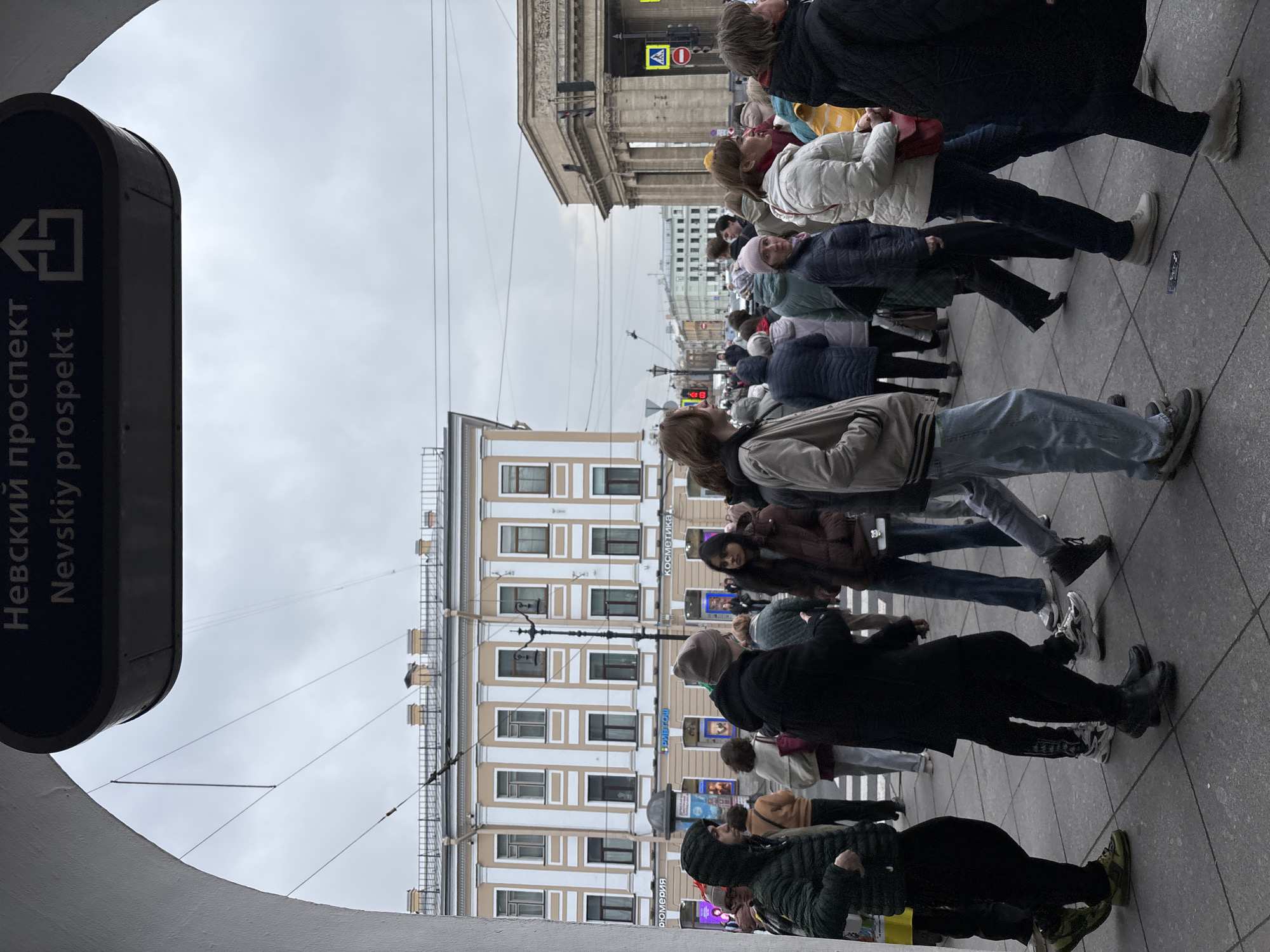 The image shows a street scene in Saint Petersburg, Russia, specifically on Nevsky Prospekt. A crowd of people is crossing the street at a pedestrian crossing. Buildings line the street, and the sky is overcast. A sign for Nevsky Prospekt is visible near what appears to be a subway entrance. The people are dressed in various types of outerwear, suggesting a cool or temperate climate.