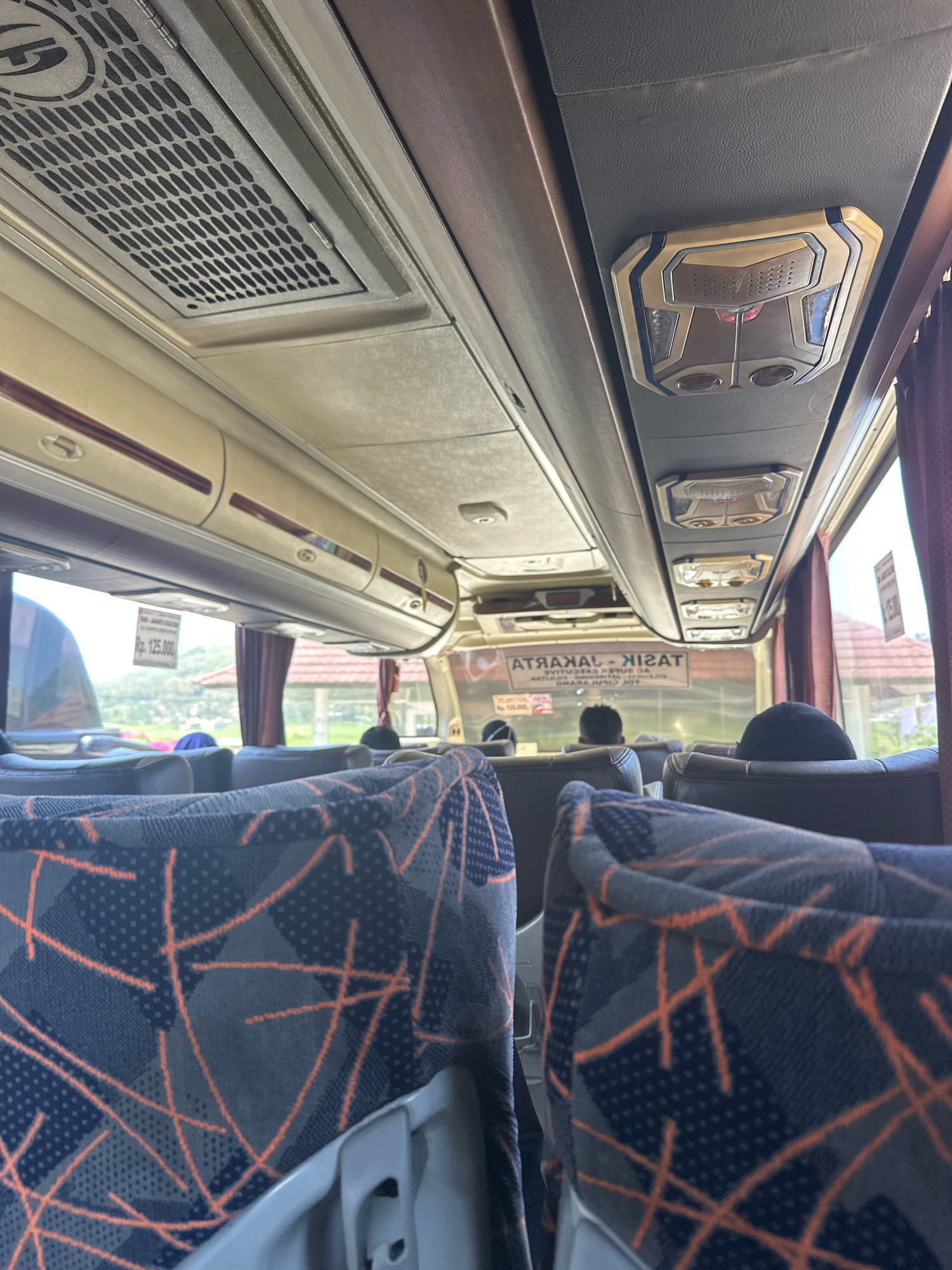The image shows the interior of a bus, viewed from the perspective of a passenger in the back. The seats in the foreground are upholstered in a dark blue fabric with a geometric pattern of orange lines and small dots. Above the seats, the bus ceiling features overhead compartments, air conditioning vents, and lighting fixtures. In the middle ground, passengers are visible sitting in their seats, facing forward. Through the windows, a bright, outdoor scene can be seen, with greenery, buildings with tiled roofs, and a glimpse of a blue sky. A sign in the window displays "Rp. 125.000" which is a price in Indonesian Rupiah. Another sign, partially visible and reversed due to being viewed from inside the bus, reads "TASIKMALAYA - GARUT". This indicates the bus is likely traveling between Tasikmalaya and Garut, or within the Garut region of Indonesia. The overall impression is of a typical intercity bus journey in Indonesia.