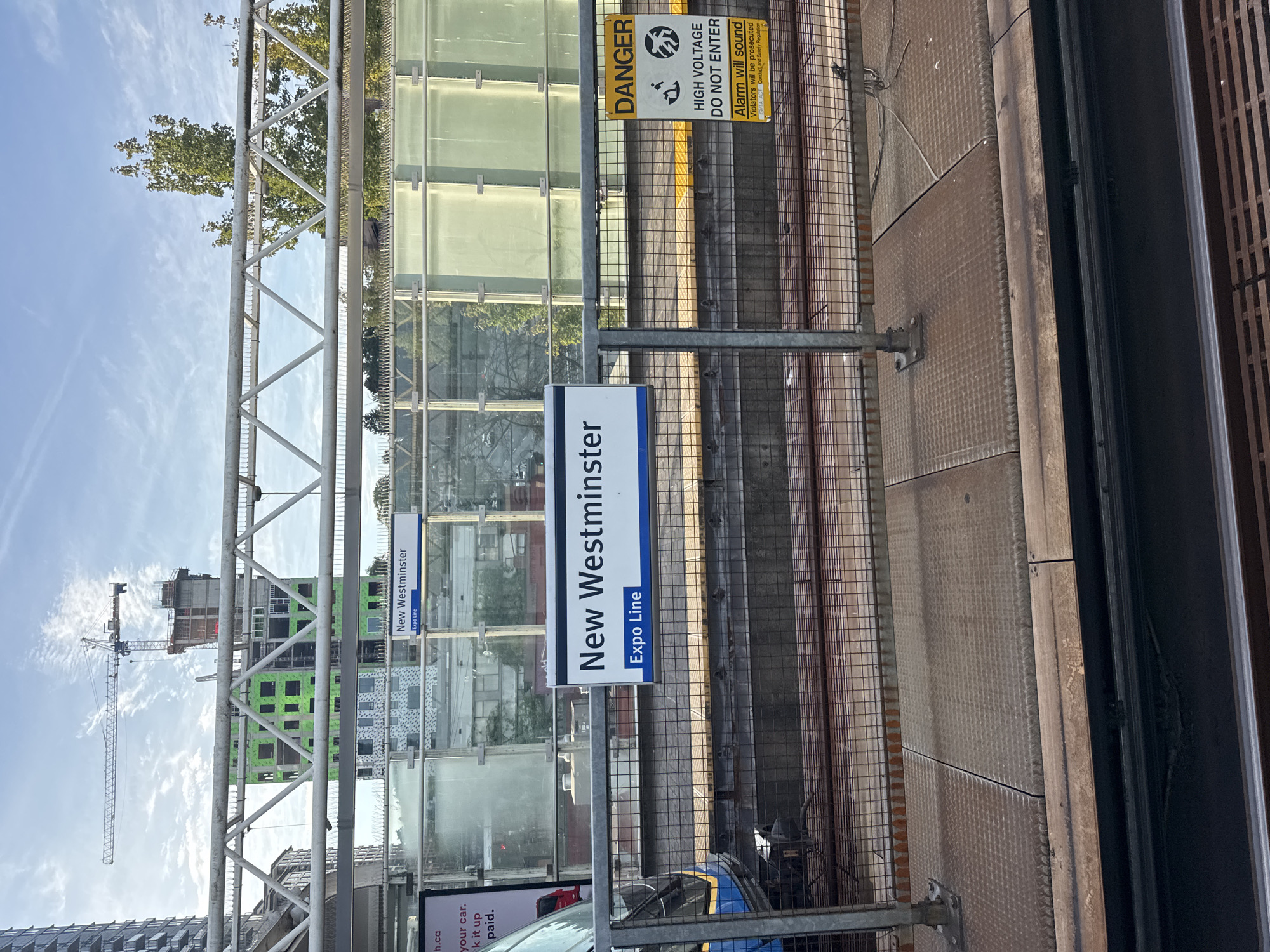 The image shows a platform at the New Westminster SkyTrain station in New Westminster, Canada. A sign on a metal railing reads "New Westminster" and "Expo Line." In the background, a train is partially visible on the tracks. To the right of the platform sign, a yellow "DANGER" sign with a high voltage warning is posted. Behind the platform, a partially constructed building with green siding is visible, along with a construction crane. The sky is blue with scattered clouds.