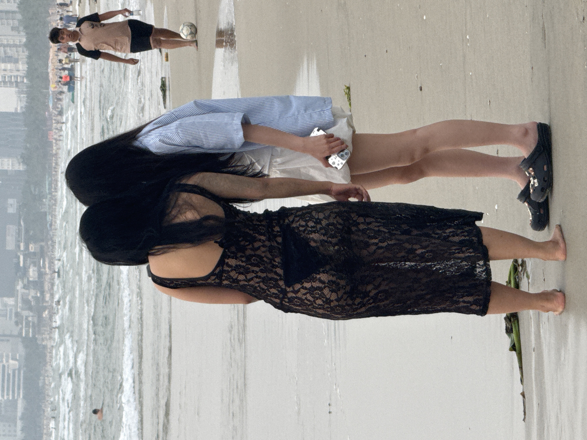 Two women stand on a beach. One wears a black lace dress, the other a striped blue shirt and white shorts. The latter holds a phone. A man in a light-colored shirt and dark shorts kicks a soccer ball in the background. The beach is sandy, and the ocean waves can be seen. Buildings are visible in the distance, suggesting this is a coastal area. Location: likely a beach in Đà Nẵng, Vietnam.