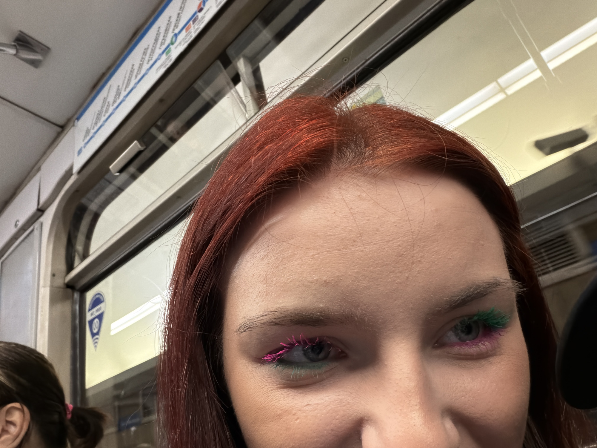 The image is a close-up, medium-shot, eye-level, indoor photograph of a person's face, specifically their eyes and forehead. The person has auburn hair and is wearing vibrant pink and green false eyelashes. The background suggests the interior of a train or subway car, with visible windows, a map or advertisement affixed to the wall above the window, and the edge of another passenger's head on the left. The lighting is artificial and appears to be from overhead fluorescent lights, creating a slightly warm cast on the scene. The focus is sharp on the subject's eyes and eyelashes, with the background slightly blurred.